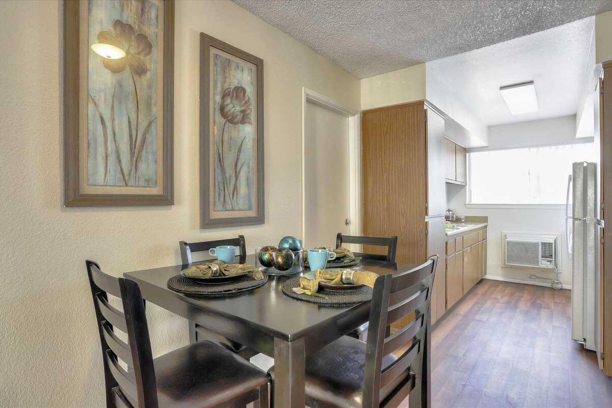 A cozy dining area featuring a table set for four with decorative plates, cups, and accessories. In the background, a well-lit kitchen with wooden cabinetry and modern appliances. Framed floral artwork adorns the walls, creating a warm and inviting atmosphere.
