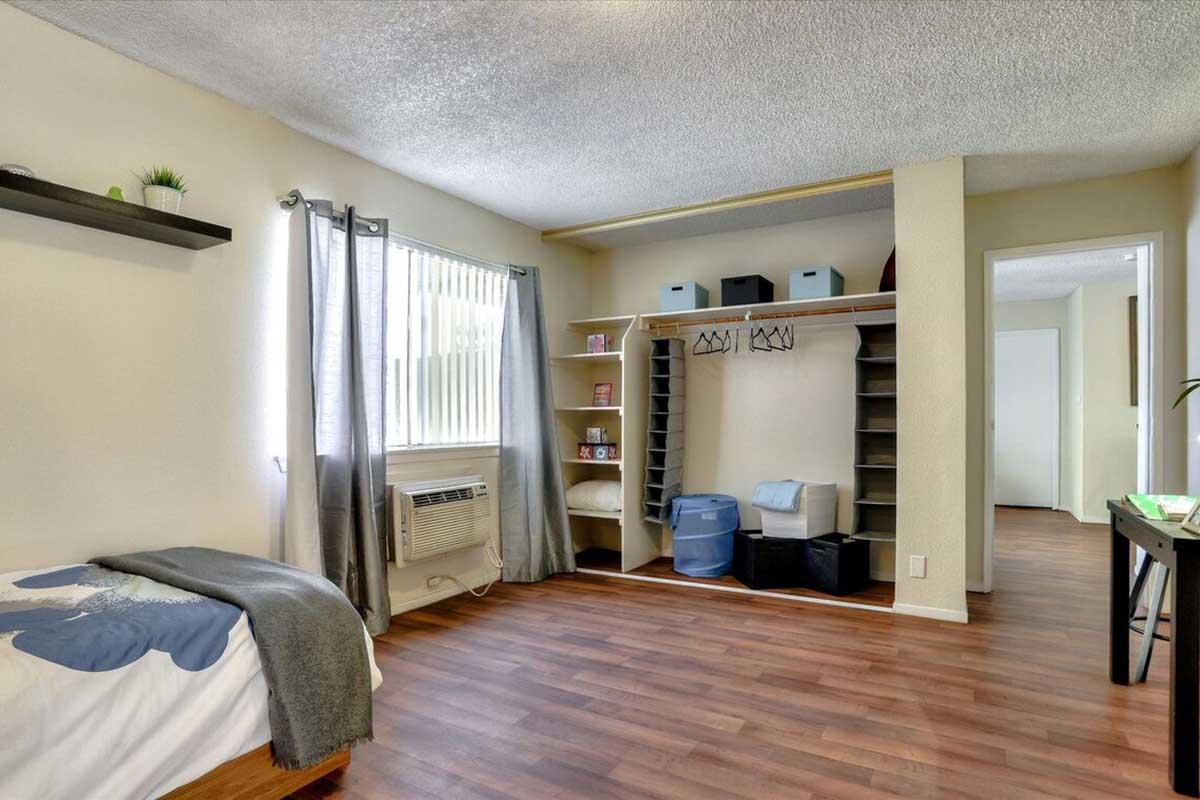 A cozy room featuring a bed with a gray blanket, air conditioning unit, large window with sheer curtains, and wooden flooring. A closet space with open shelves, storage bins, and hanging rods is visible, creating a functional and organized atmosphere.