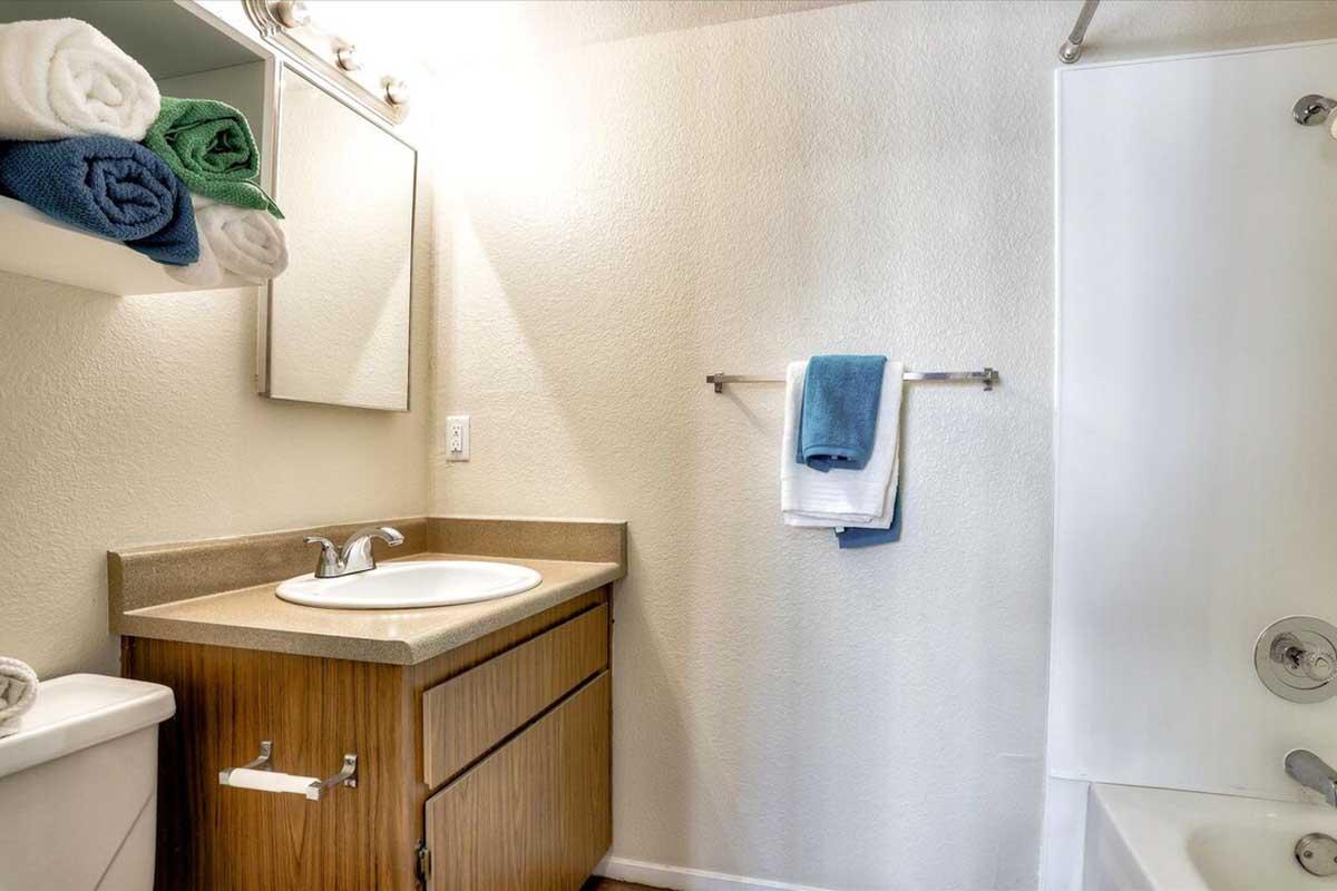 A clean bathroom featuring a sink with a wooden cabinet, a mirror, and towels neatly arranged on a shelf. The bathtub is equipped with a shower, and there are additional towels hanging on a rack. The walls are painted a light color, contributing to a bright and organized appearance.
