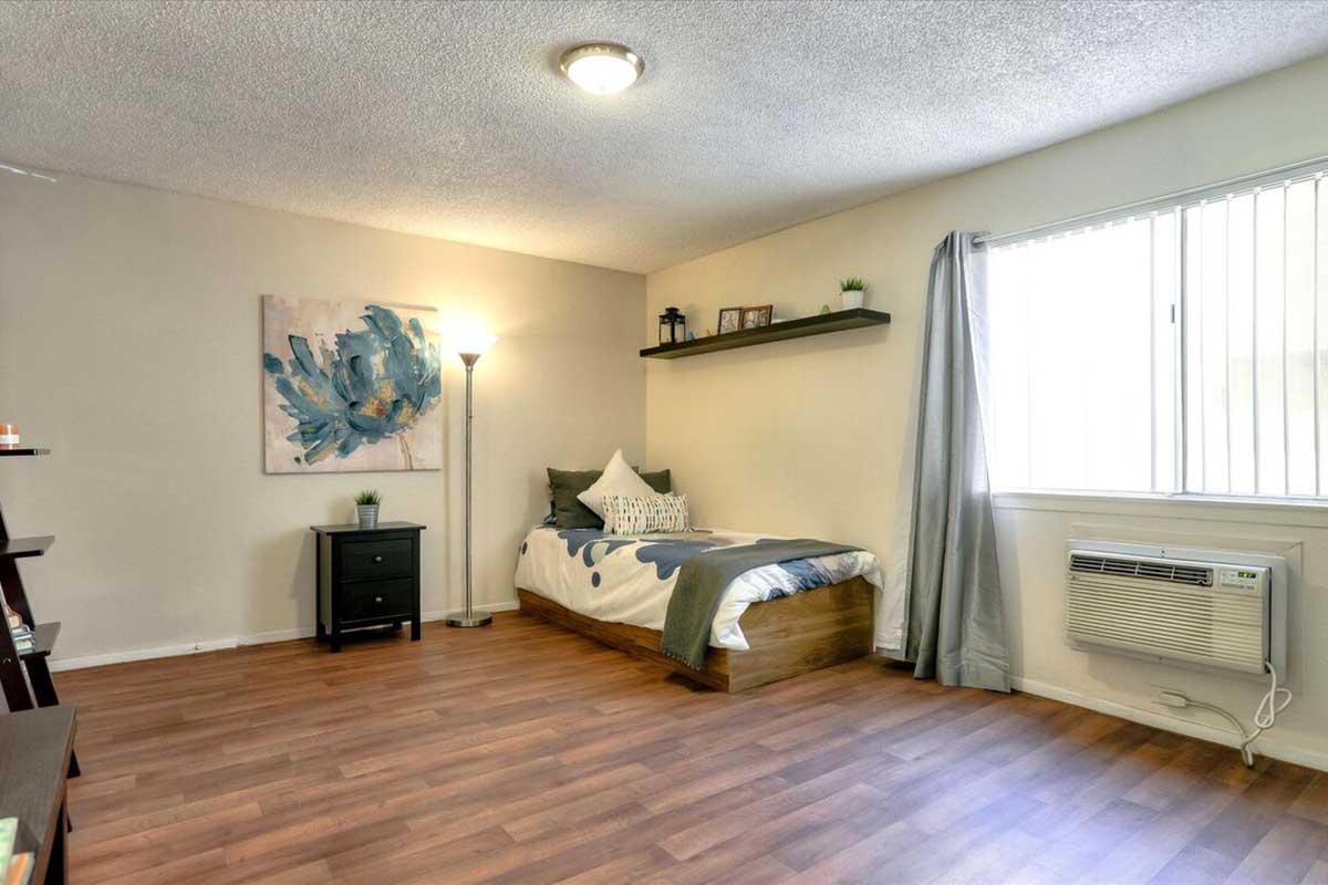A cozy bedroom featuring a single bed with decorative pillows, a nightstand beside it, and a floor lamp. A shelf with small decor items is mounted on the wall above the bed. Large window with white blinds allows natural light in, and an air conditioning unit is visible below the window. The flooring is a warm wood tone.