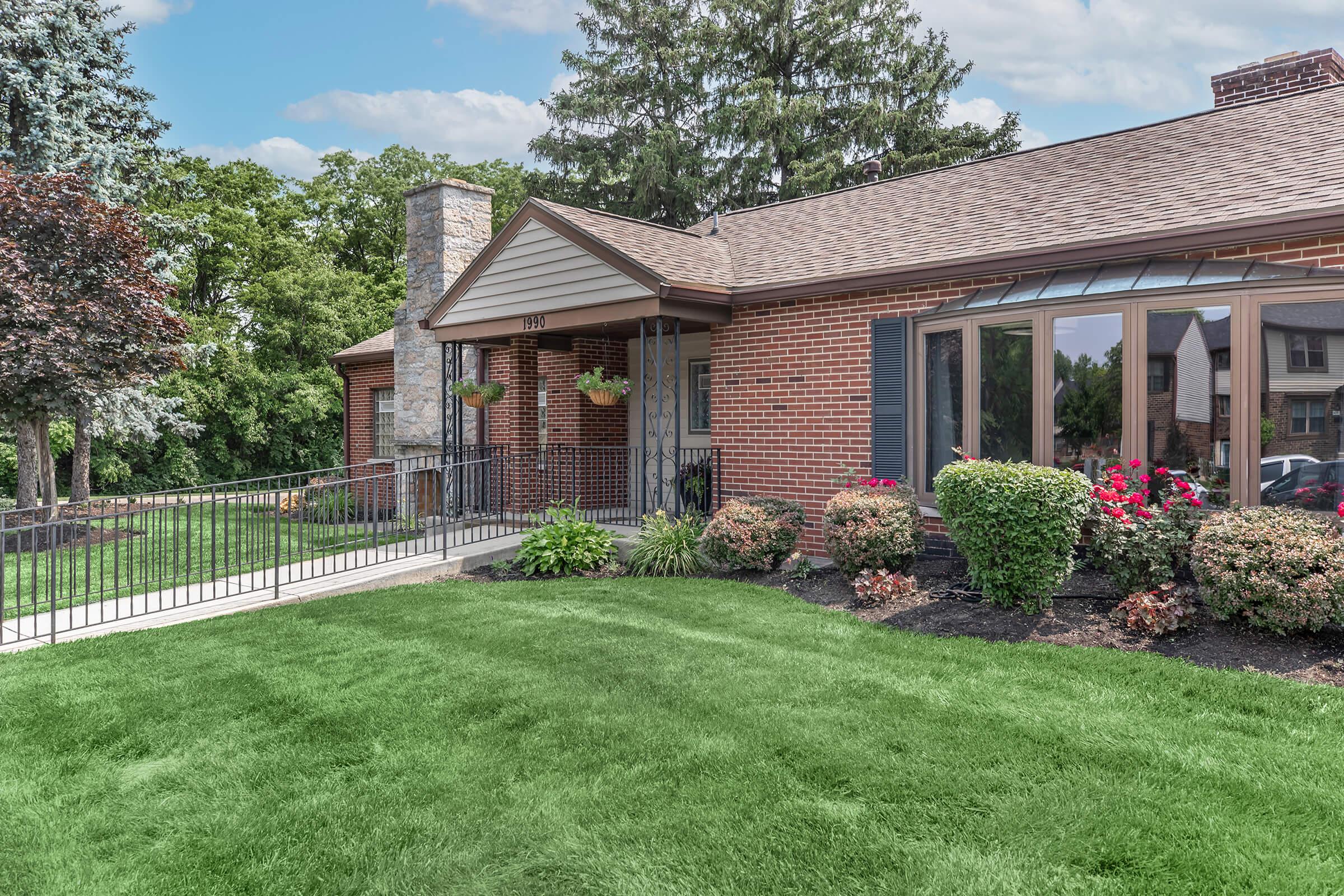 A well-maintained brick house with a sloped roof, surrounded by lush green lawns and manicured flower beds. The front features large windows, planters, and a stone chimney, with a pathway leading to the entrance. Trees and shrubs add to the pleasant landscaping on a sunny day.