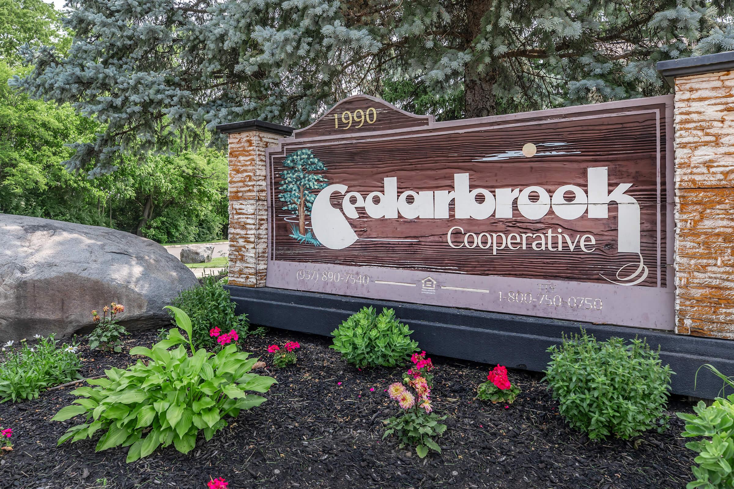 Sign for Cedarbrook Cooperative, established in 1990, featuring a wooden design with green trees. The sign includes contact information and is surrounded by colorful flowers and greenery, set against a backdrop of evergreen trees.