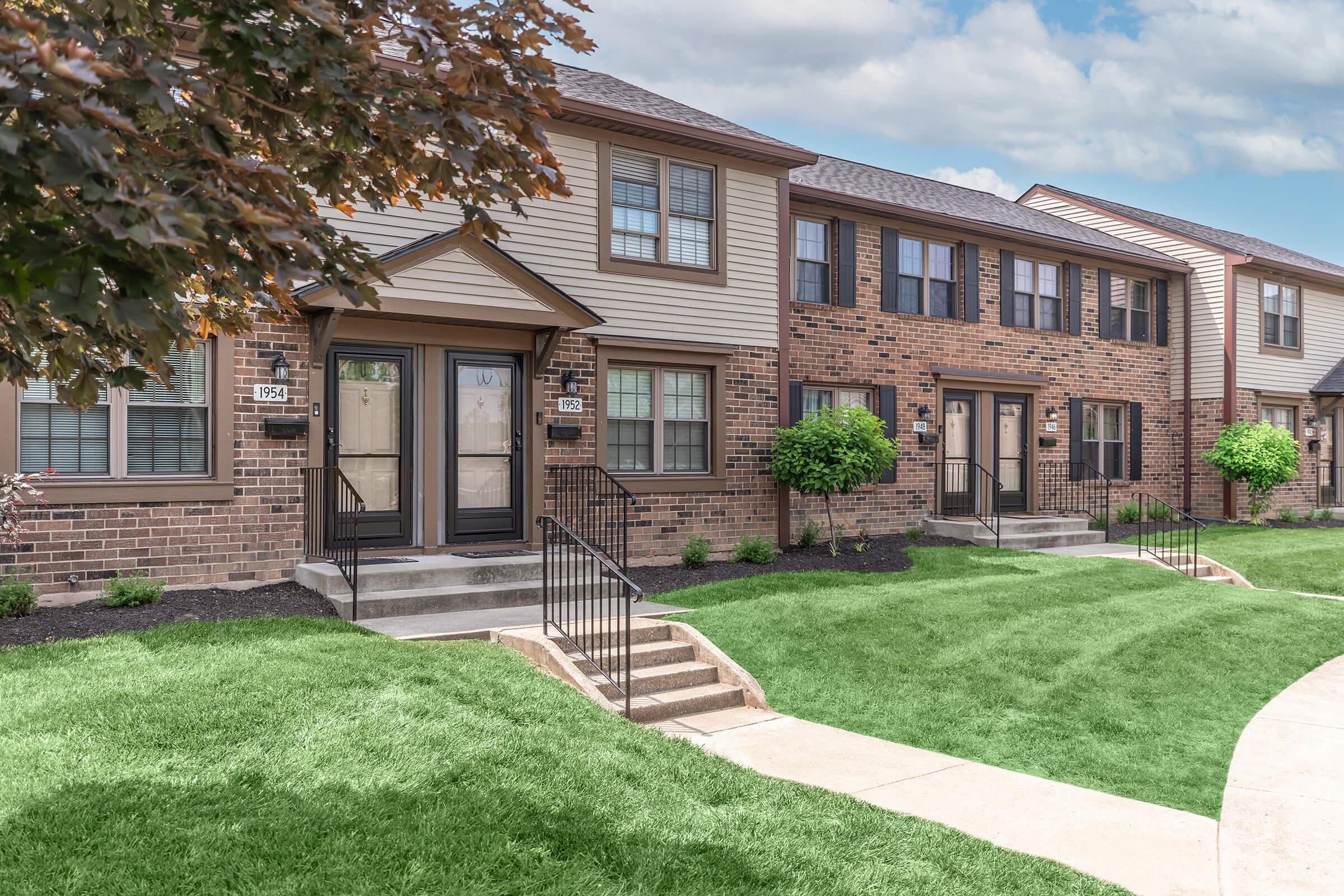 Two-story residential buildings with a mix of brick and wood siding. Each unit has its own entrance with black railings leading to the door. Lush green lawns and small trees are visible in the front yard, and a concrete pathway runs alongside the buildings. The sky is partly cloudy.