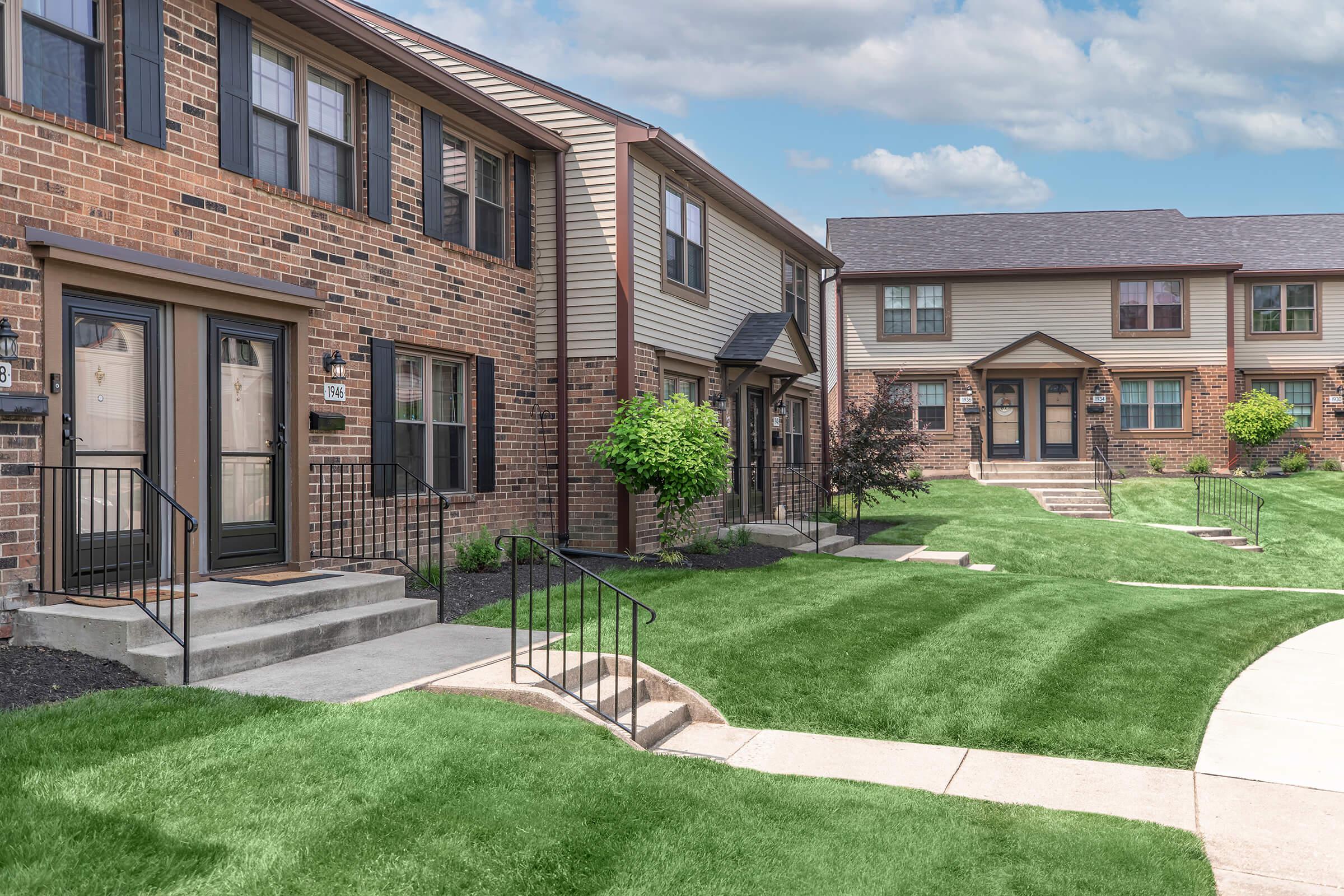 A modern apartment complex with brick and beige siding. The buildings have multiple entrances with stairs leading up to them, surrounded by well-maintained green lawns and pathways. There are small trees and shrubs adding to the landscape, under a partly cloudy sky.