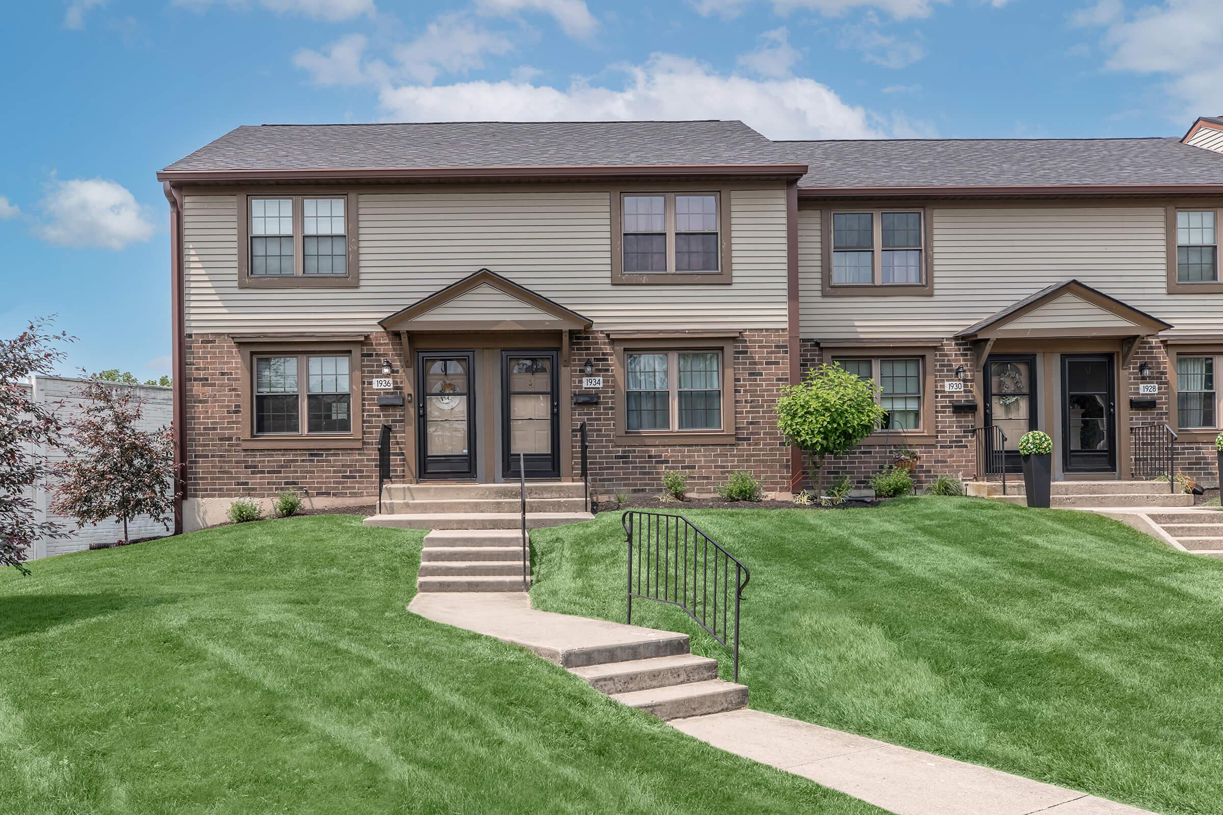 A well-maintained two-story townhouse with a brick and siding exterior, featuring a green lawn and neatly manicured bushes. A sidewalk leads to the front doors, and the entrance has steps and railings. The sky is clear with a few clouds in the background.
