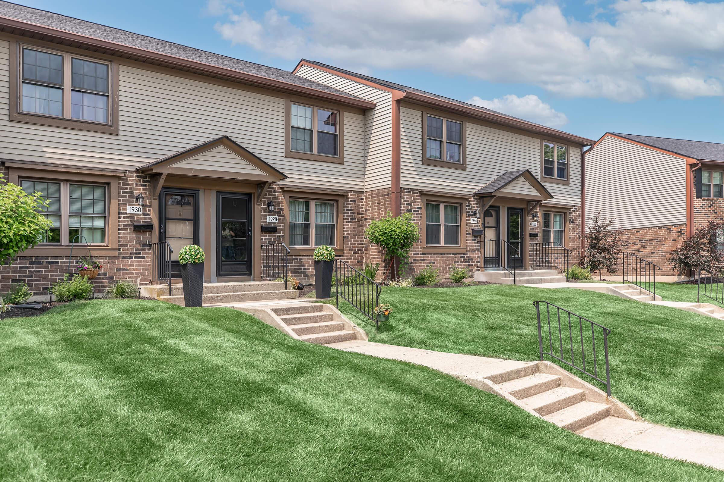 A row of two-story townhouses with brown siding and stone accents. Each unit features front steps, small gardens, and planters. The well-manicured green lawn complements the modern architecture under a blue sky with light clouds.