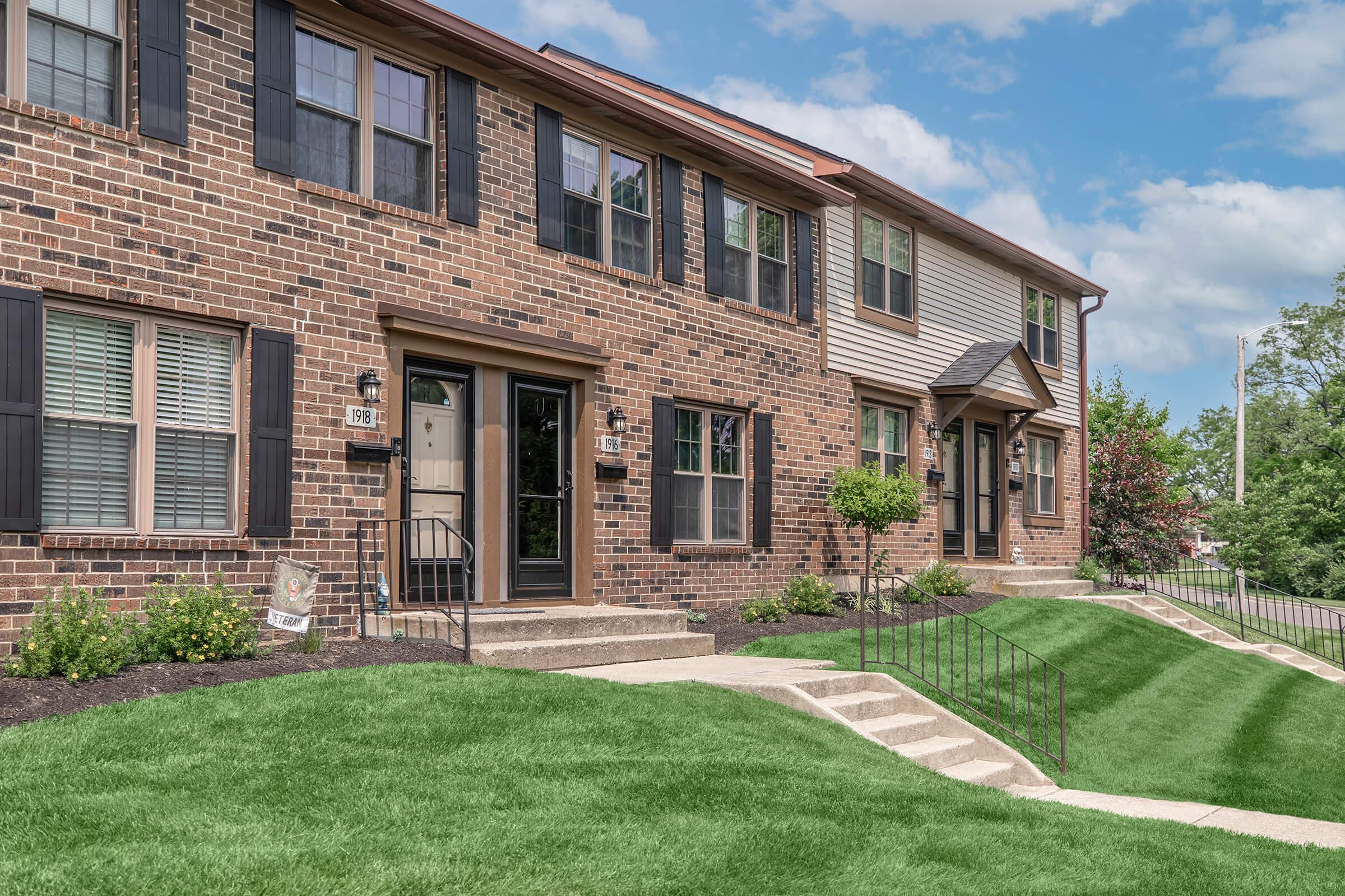 A row of modern townhouses with brick facades and black shutters. Each unit has a porch with steps leading up to the door. The landscaped lawn is green and well-maintained, with small bushes and a sidewalk. A clear blue sky is visible in the background, suggesting a pleasant day.