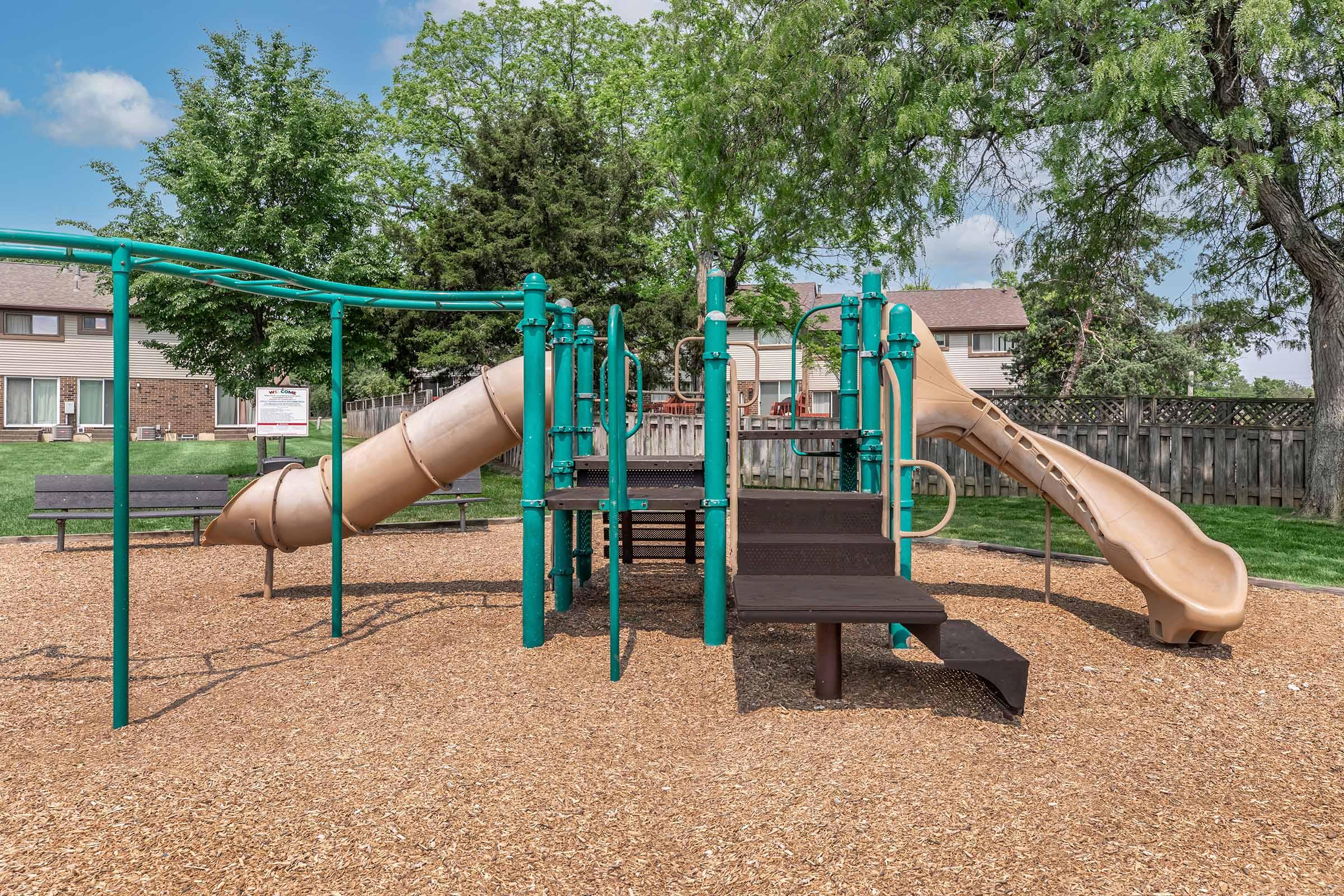 A playground featuring a tan slide, climbing structures, and a series of steps, surrounded by wood chips. Green equipment is prominent, with benches nearby. Lush green trees and a residential area can be seen in the background, providing a serene environment for children to play.