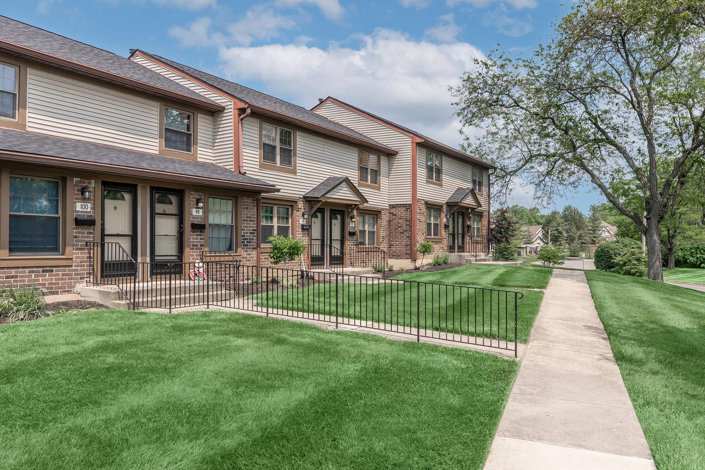A row of townhouse-style buildings with brick and siding exteriors. Each unit features a front porch and is surrounded by well-maintained lawns. A paved walkway runs alongside the homes, and a green tree is visible nearby under a partly cloudy sky.