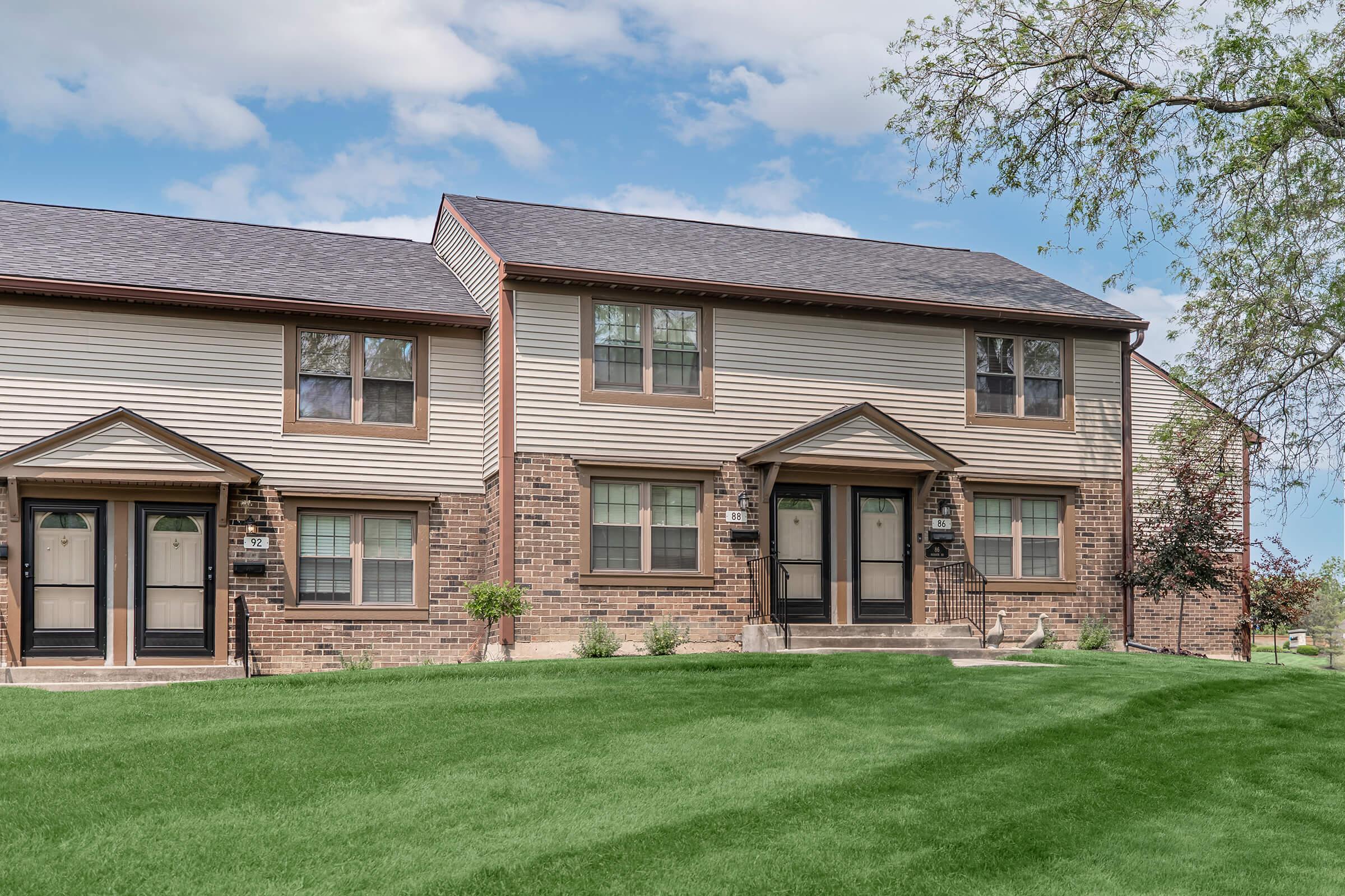 A two-story brick and siding townhouse with a well-manicured lawn. The building features multiple entrances, small shrubs, and a clear blue sky in the background.