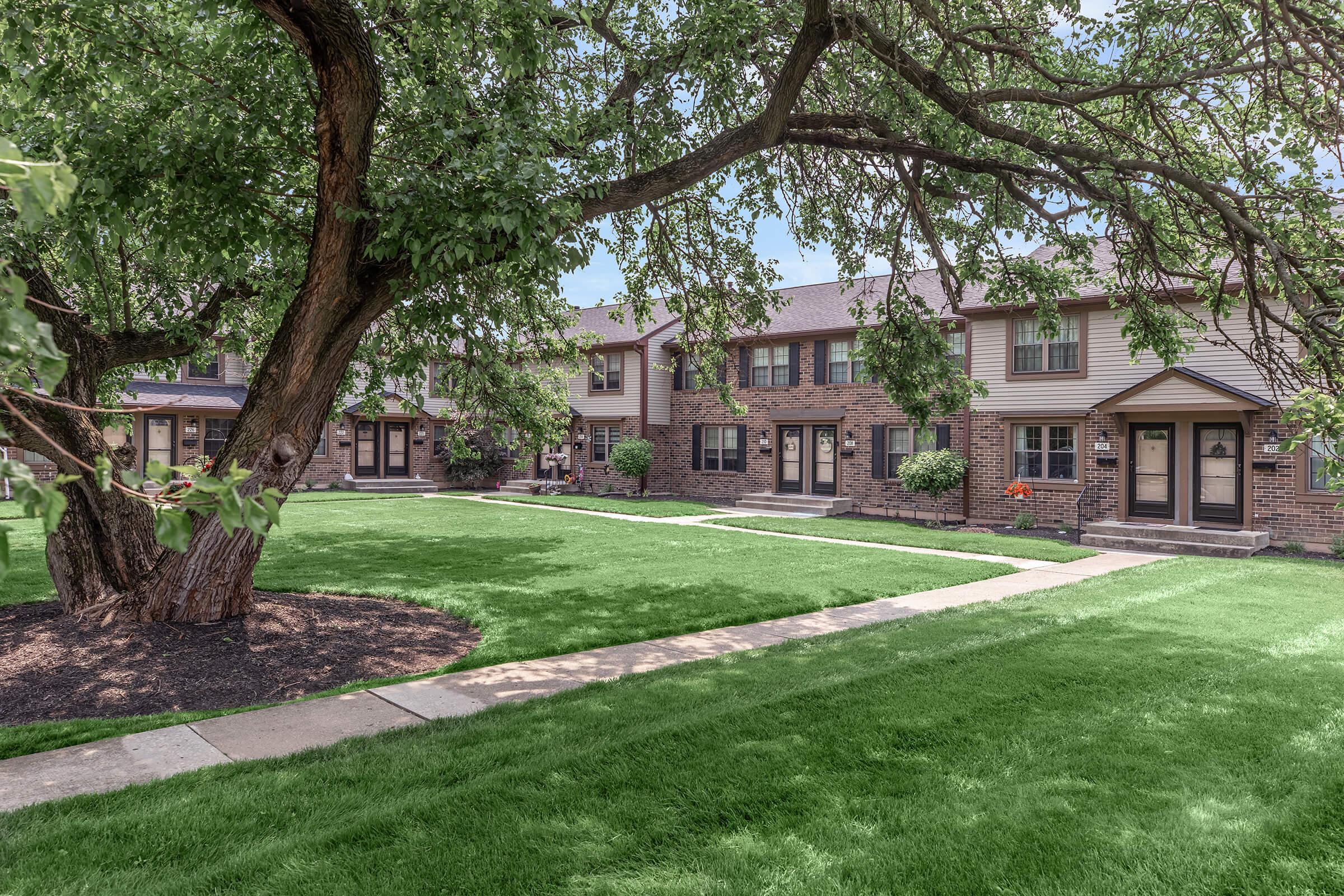 A well-maintained residential complex featuring brick and siding townhouses arranged around a green lawn. Mature trees provide shade, and pathways lead between the buildings. Flower beds add color to the inviting outdoor space. The scene conveys a peaceful and welcoming neighborhood atmosphere.