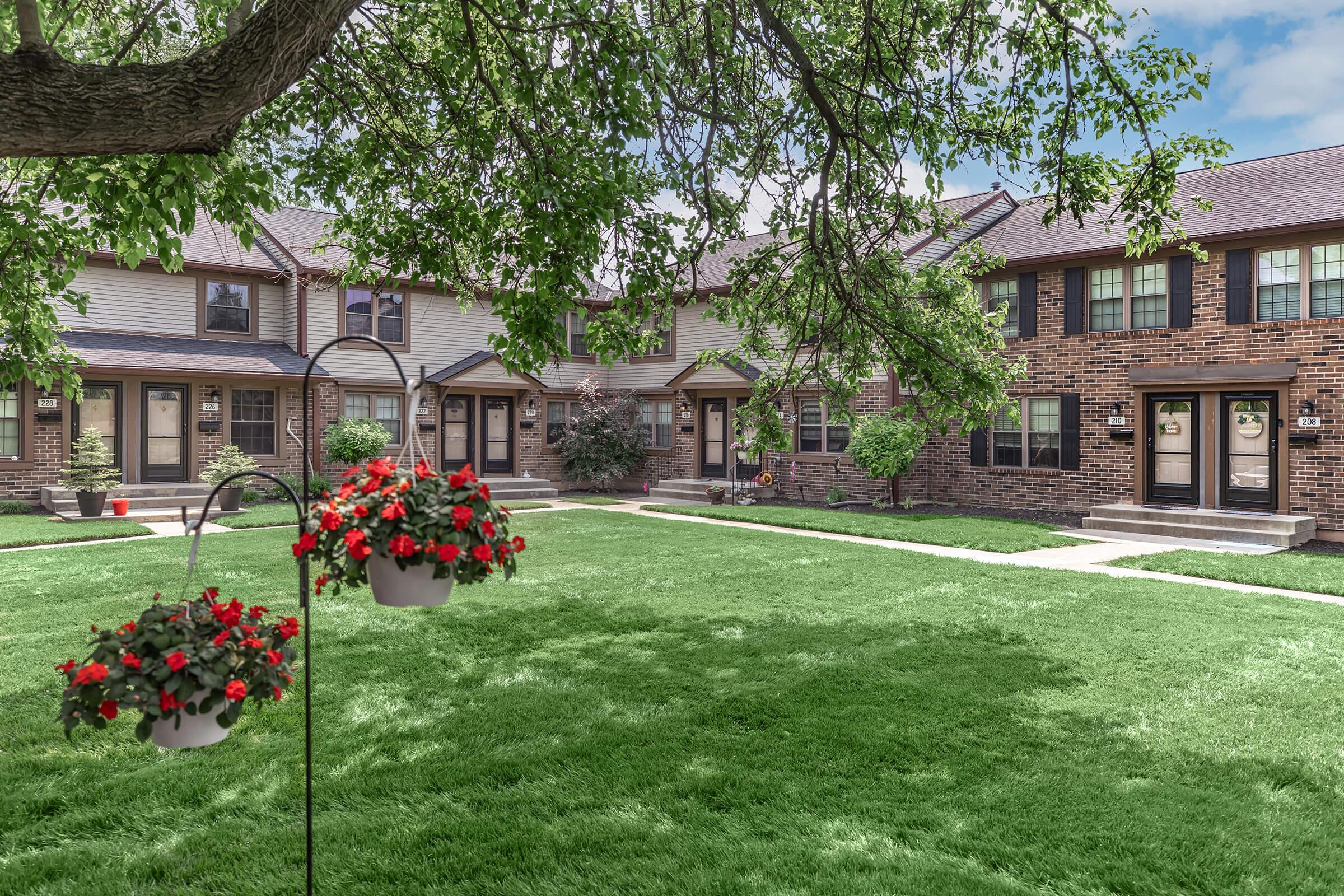 A well-maintained green courtyard featuring a grassy area and flower baskets hanging from a tree. The background shows two rows of townhouses, with a mix of brick and siding exteriors. The scene is bright and inviting, reflecting a suburban residential setting.