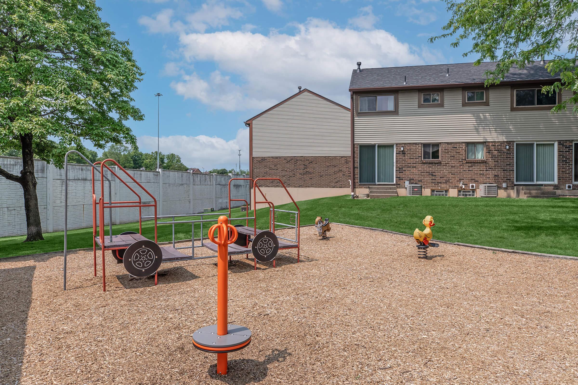 A residential playground featuring colorful play structures on a gravel surface. Two children are playing: one is near a spinning device, while the other is on a seesaw. The background shows a grassy area and two-story townhouses under a partly cloudy sky.