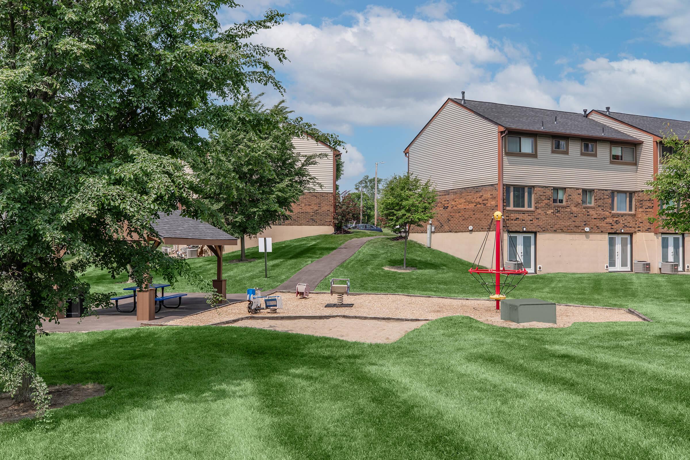 A park area featuring a playground with a swing set and a small slide, surrounded by well-maintained grass. Behind the playground, there are several residential buildings with a pathway leading between them. The scene is set under a partly cloudy sky, with trees providing some shade.