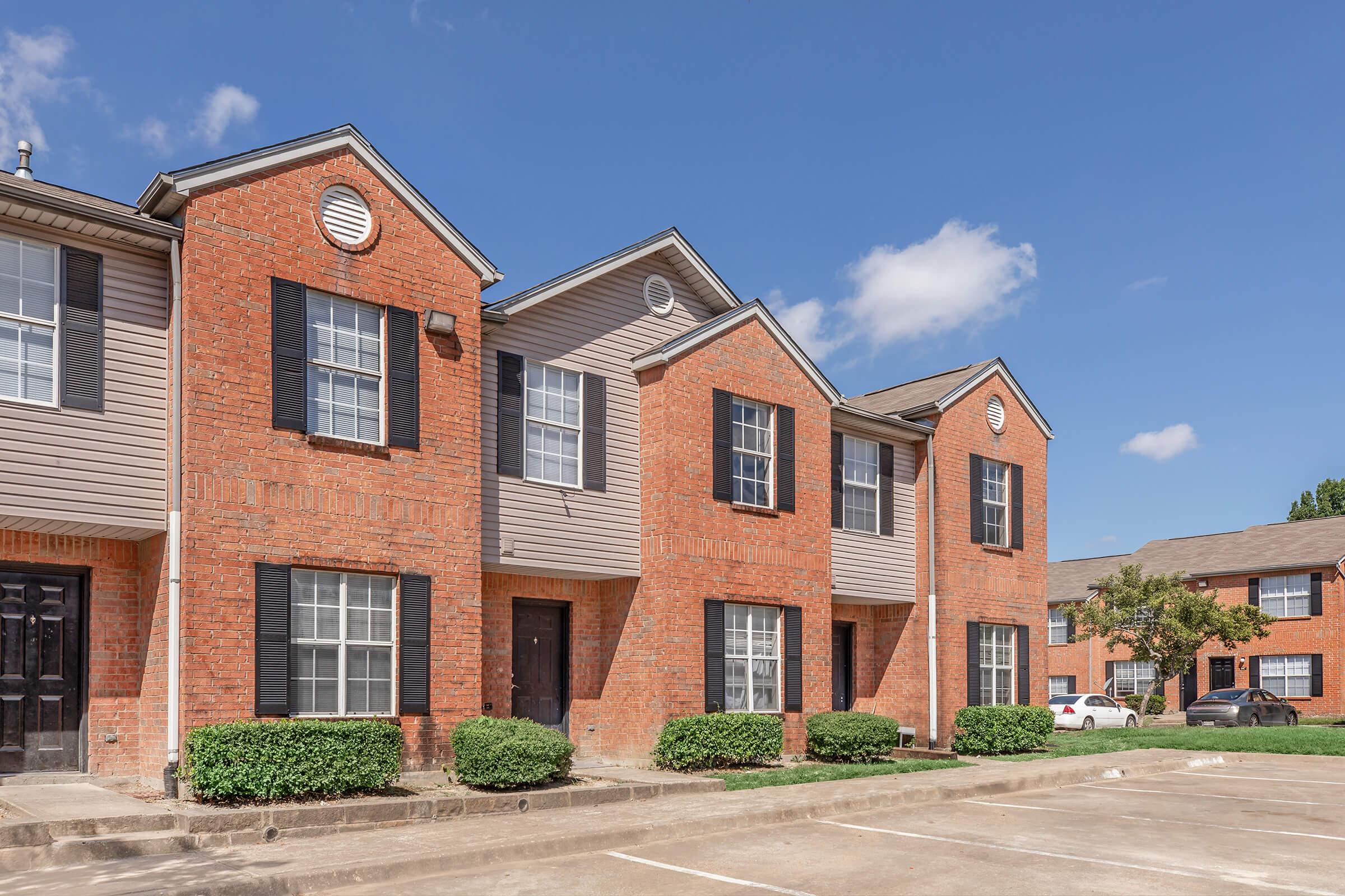 A row of brick and siding apartment buildings under a clear blue sky. The buildings have multiple windows with black shutters and a neatly landscaped area in front. There are parking spots available nearby, adding to the residential setting.