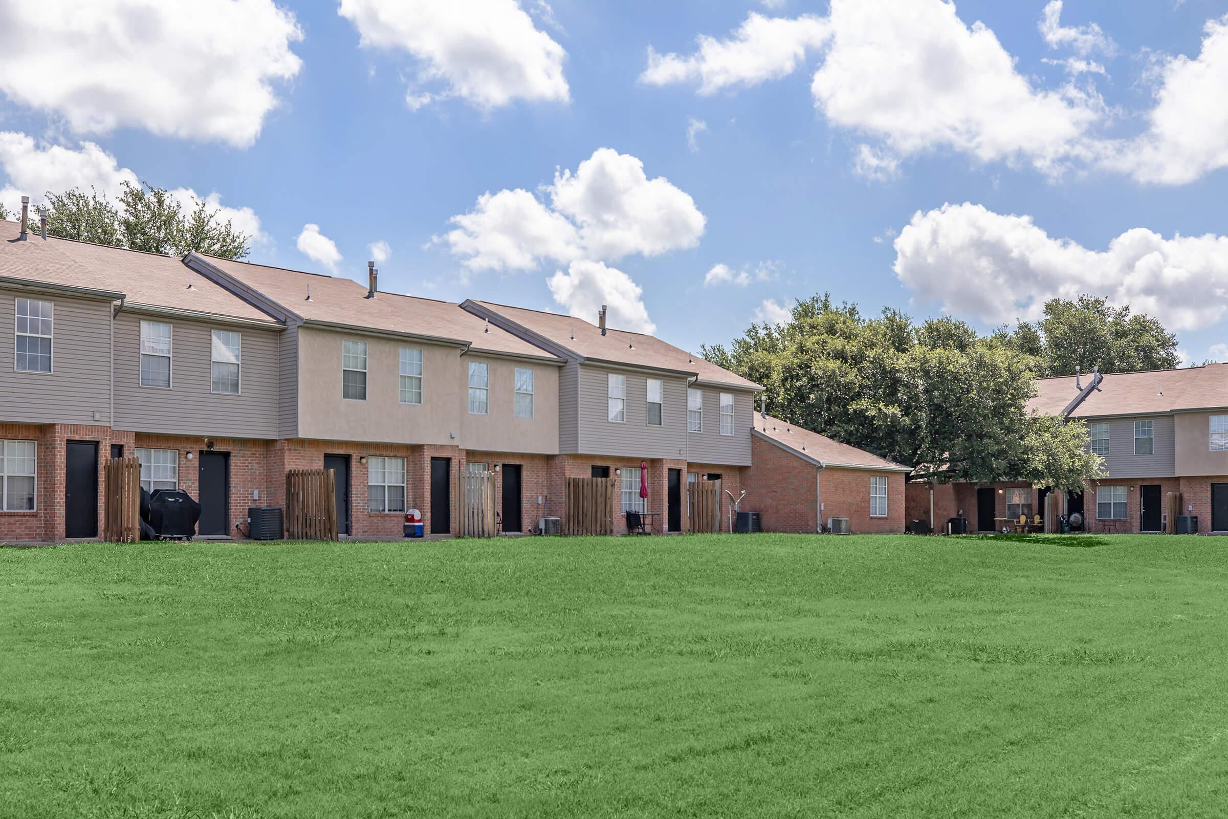 A row of residential buildings with a grassy lawn in the foreground. The buildings are two-story, featuring beige and red brick facades, with porches and individual entrances. The sky is bright blue with fluffy white clouds, creating a pleasant suburban scene.