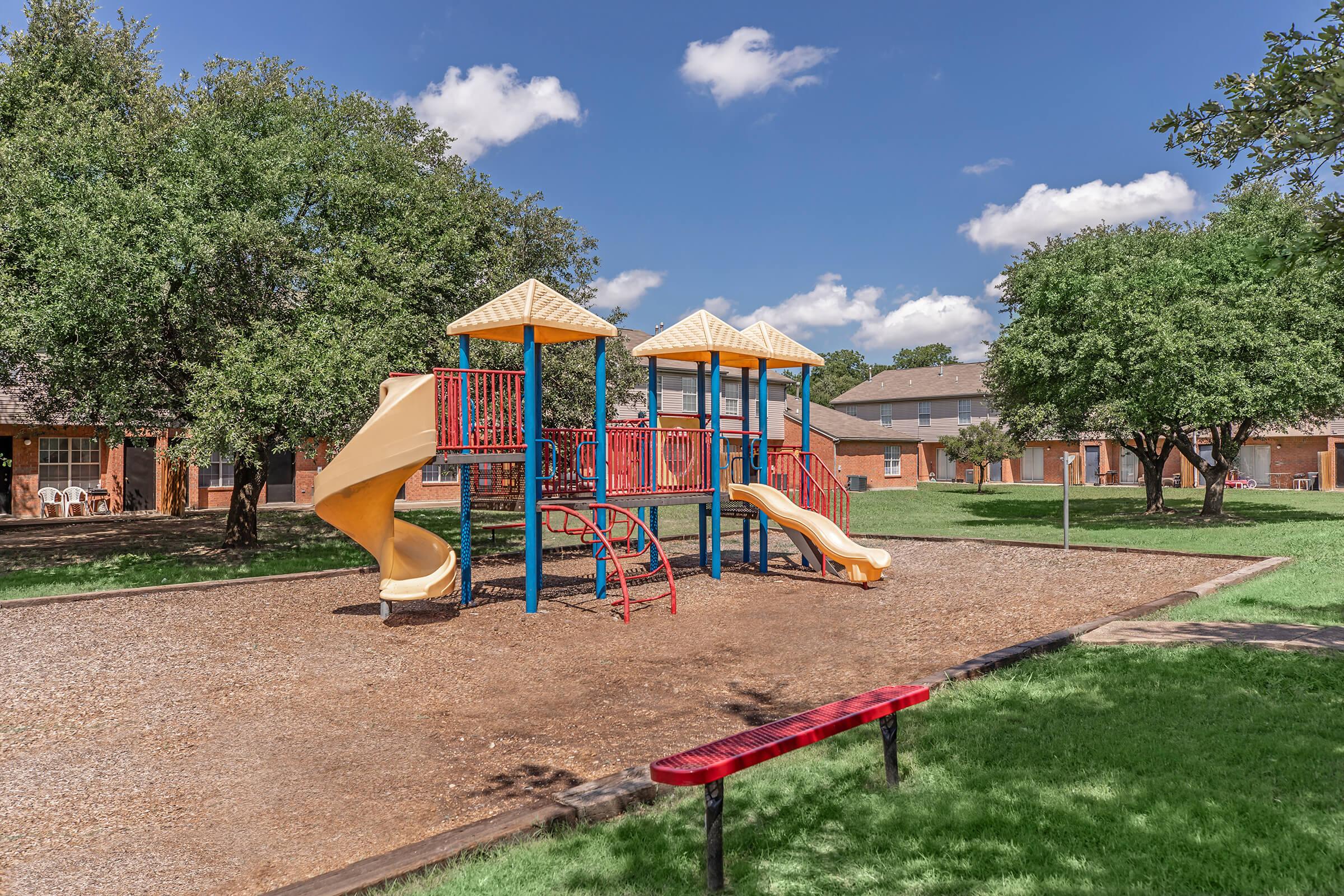A colorful playground with a spiral slide and two slides, set on a gravel surface surrounded by green grass. Nearby are trees and a red bench, with buildings in the background under a blue sky with fluffy white clouds.