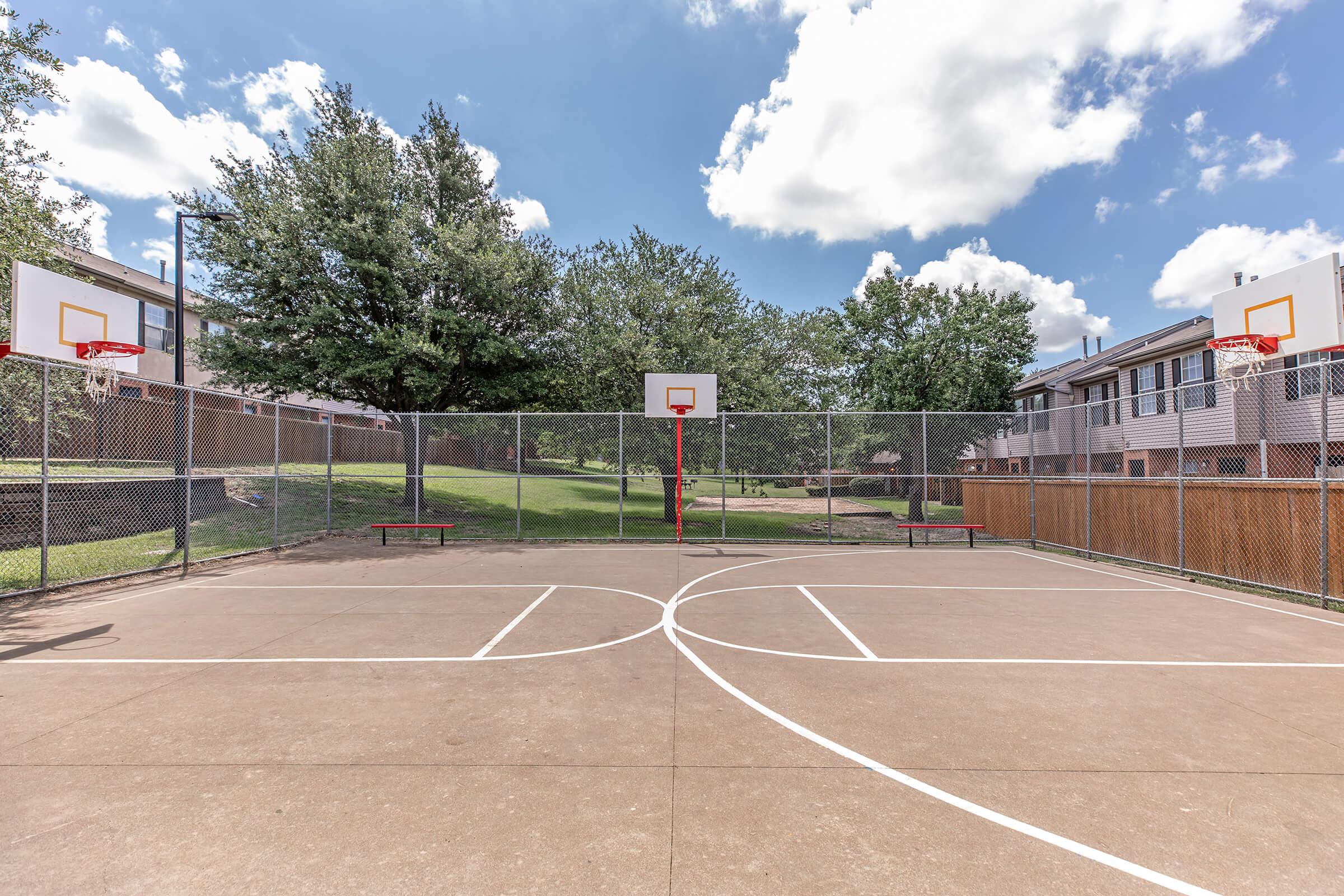 A well-maintained outdoor basketball court featuring two hoops, with clear blue skies and fluffy clouds above. Surrounding the court are trees and residential buildings, enclosed by a chain-link fence. The court surface is marked with white lines, indicating play areas.