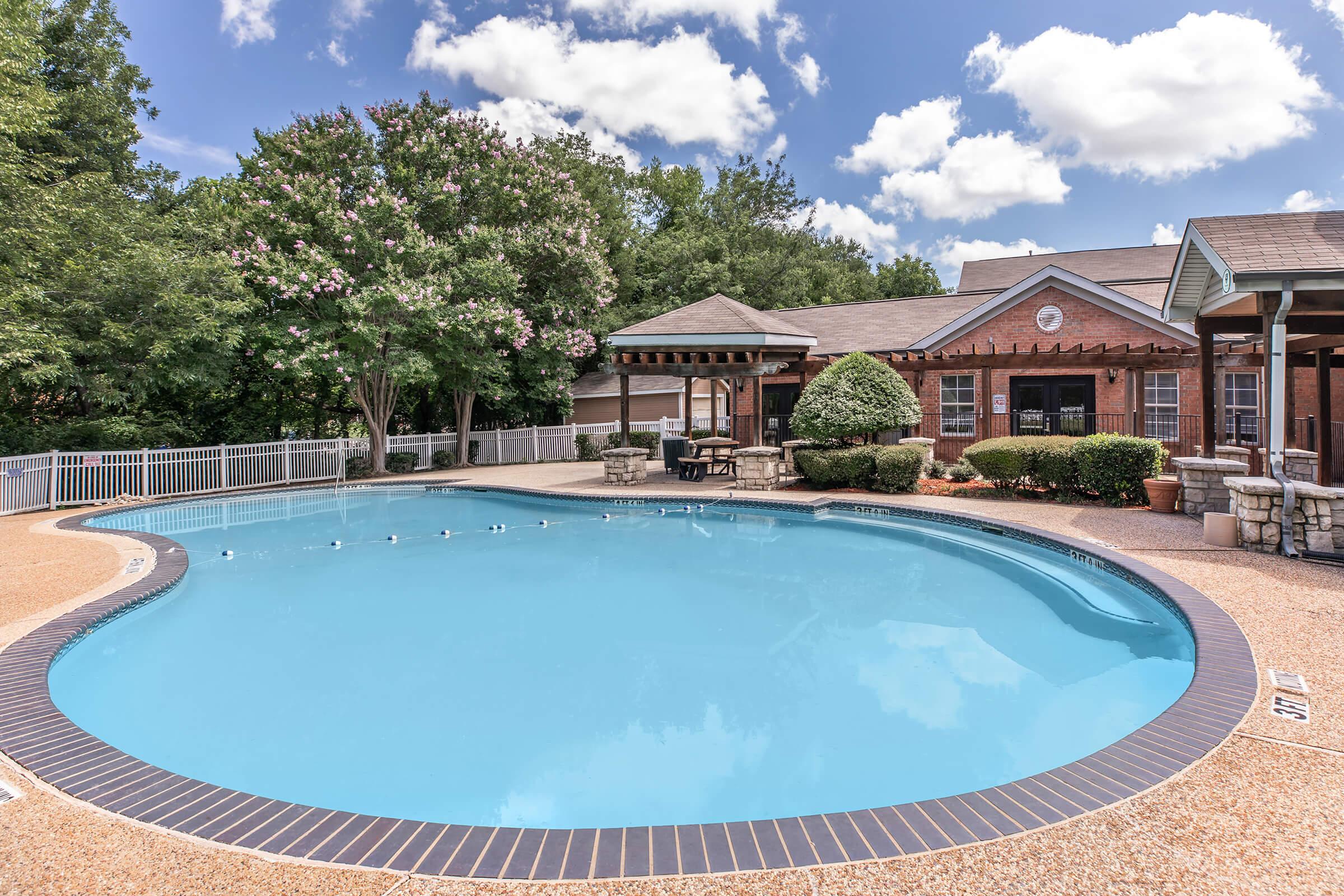 A tranquil swimming pool surrounded by well-manicured landscaping, featuring lush green trees and flowering shrubs. The pool is oval-shaped with a blue water surface and bordered by a tan deck. Adjacent is a covered seating area and a brick building, under a bright blue sky with scattered clouds.