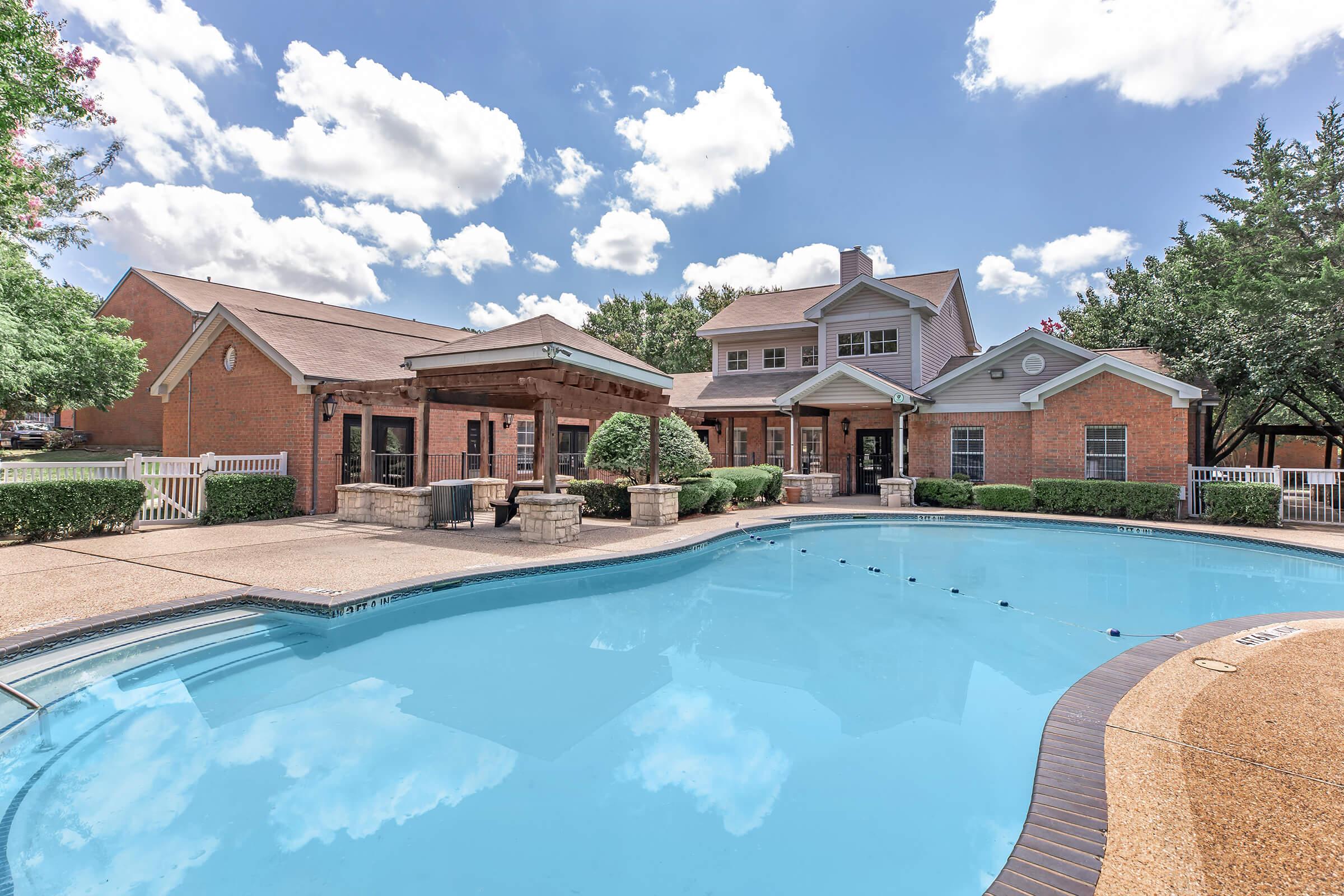 A beautiful outdoor area featuring a swimming pool surrounded by landscaped greenery, a stone patio, and a brick building in the background. Fluffy clouds fill the blue sky, creating a sunny and inviting atmosphere perfect for relaxation and leisure.