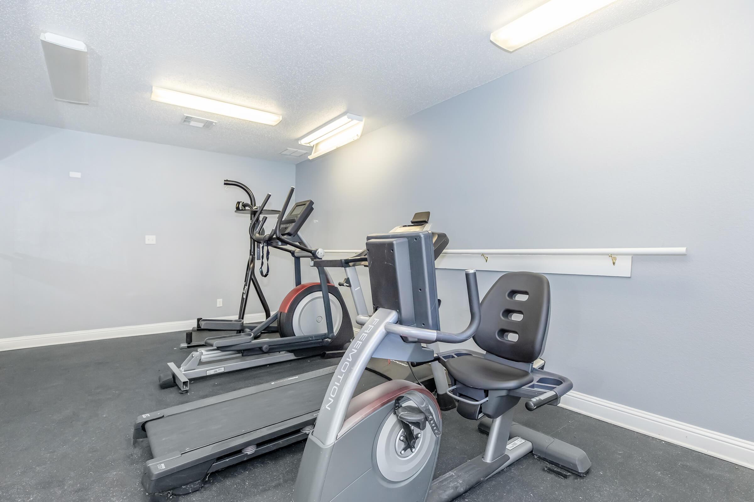A small, well-lit gym space featuring two pieces of exercise equipment: an elliptical and a stationary bike. The walls are painted light blue, and there is a wall-mounted bar for support or stretching. The floor is dark, and the overall atmosphere is clean and functional.