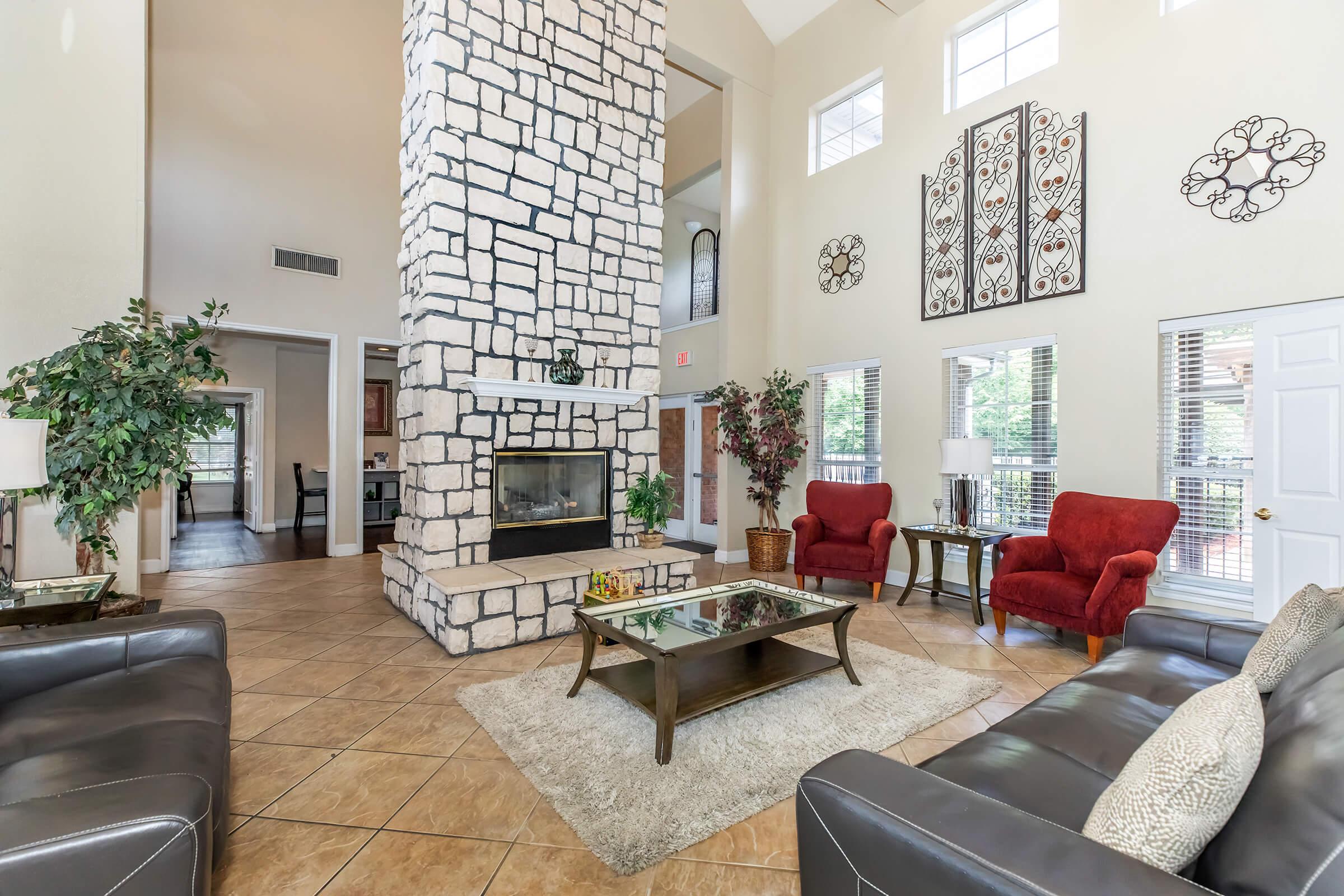 A spacious living room featuring a tall stone fireplace as a focal point, with large windows allowing natural light. The room includes two red armchairs, a coffee table on a light rug, dark leather sofas, and decorative plants, creating a cozy and inviting atmosphere.