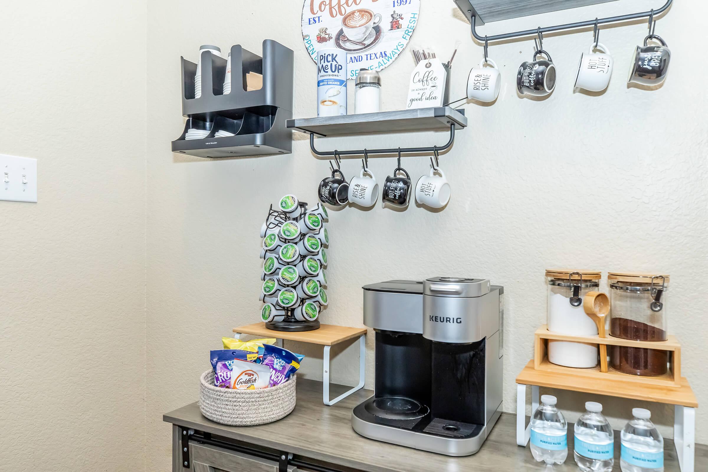 A cozy coffee station featuring a Keurig coffee maker, a variety of K-cup pods organized in a vertical holder, ceramic mugs on a rack, and supplies like sugar, creamer, and bottled water. Wall decor includes a playful sign about coffee. The setup is on a wooden countertop against a light-colored wall.