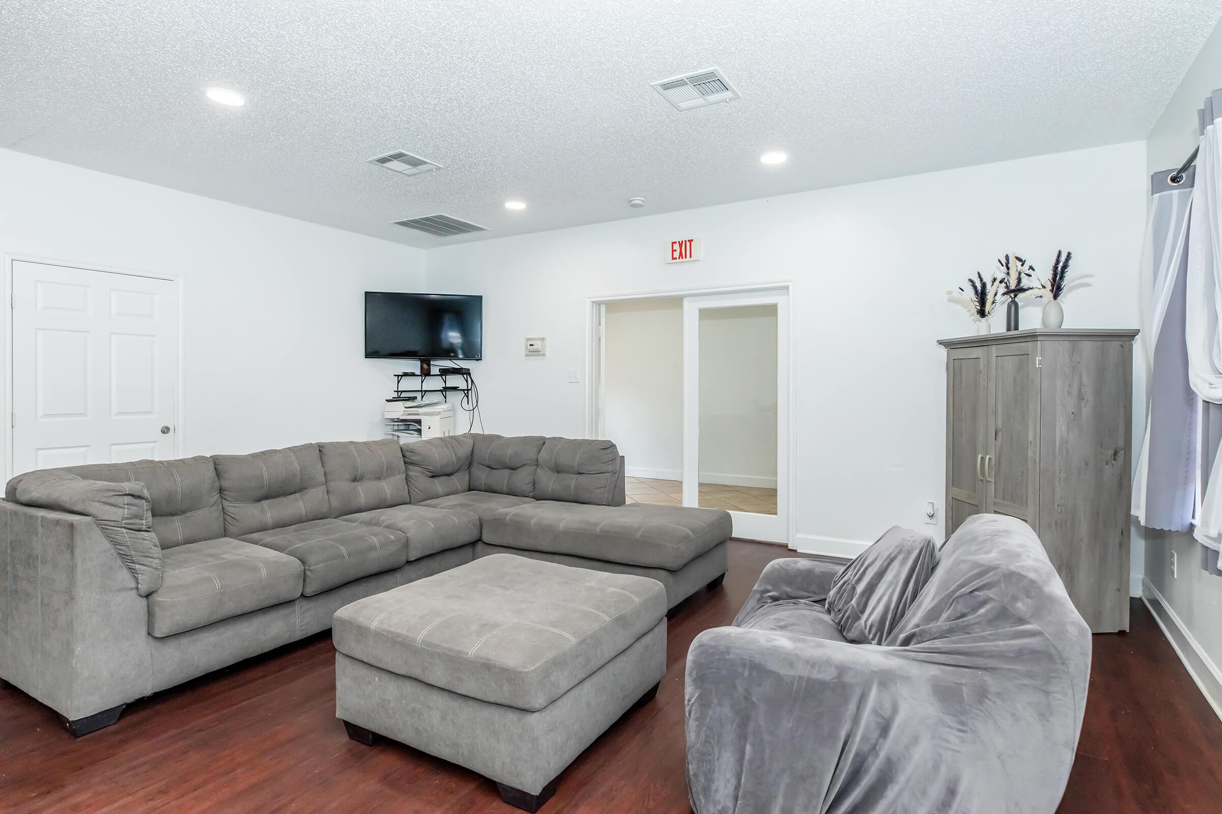 A modern living room featuring a large gray sectional sofa and an ottoman, a flat-screen TV mounted on the wall, and a gray armchair. The walls are painted white, and there is a door leading to another room, with soft lighting creating a cozy atmosphere.