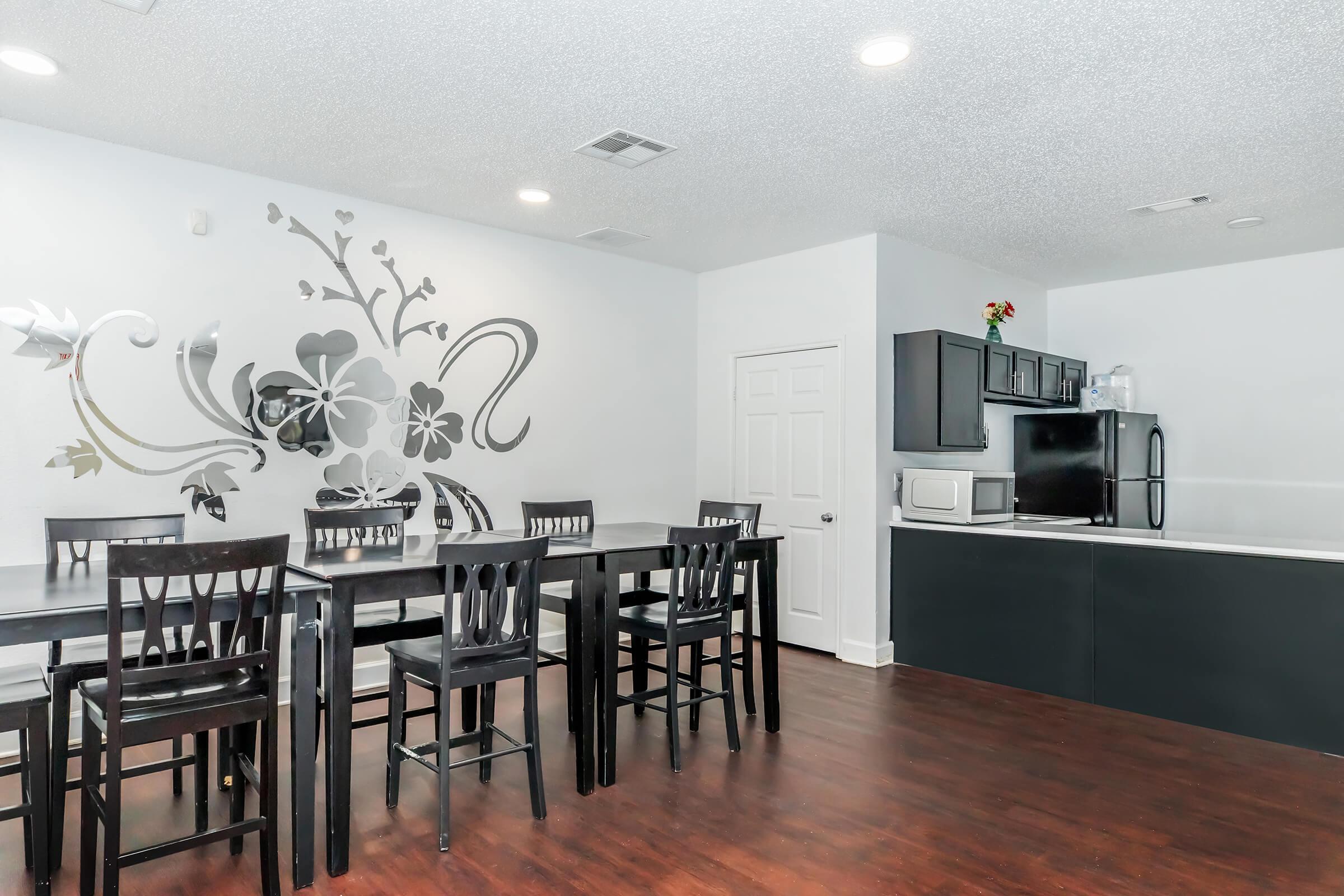 Modern kitchen dining area featuring a wooden table with black chairs, a decorative floral wall decal, and sleek black cabinetry. The kitchen includes a microwave and refrigerator against a white wall, with natural light illuminating the space.