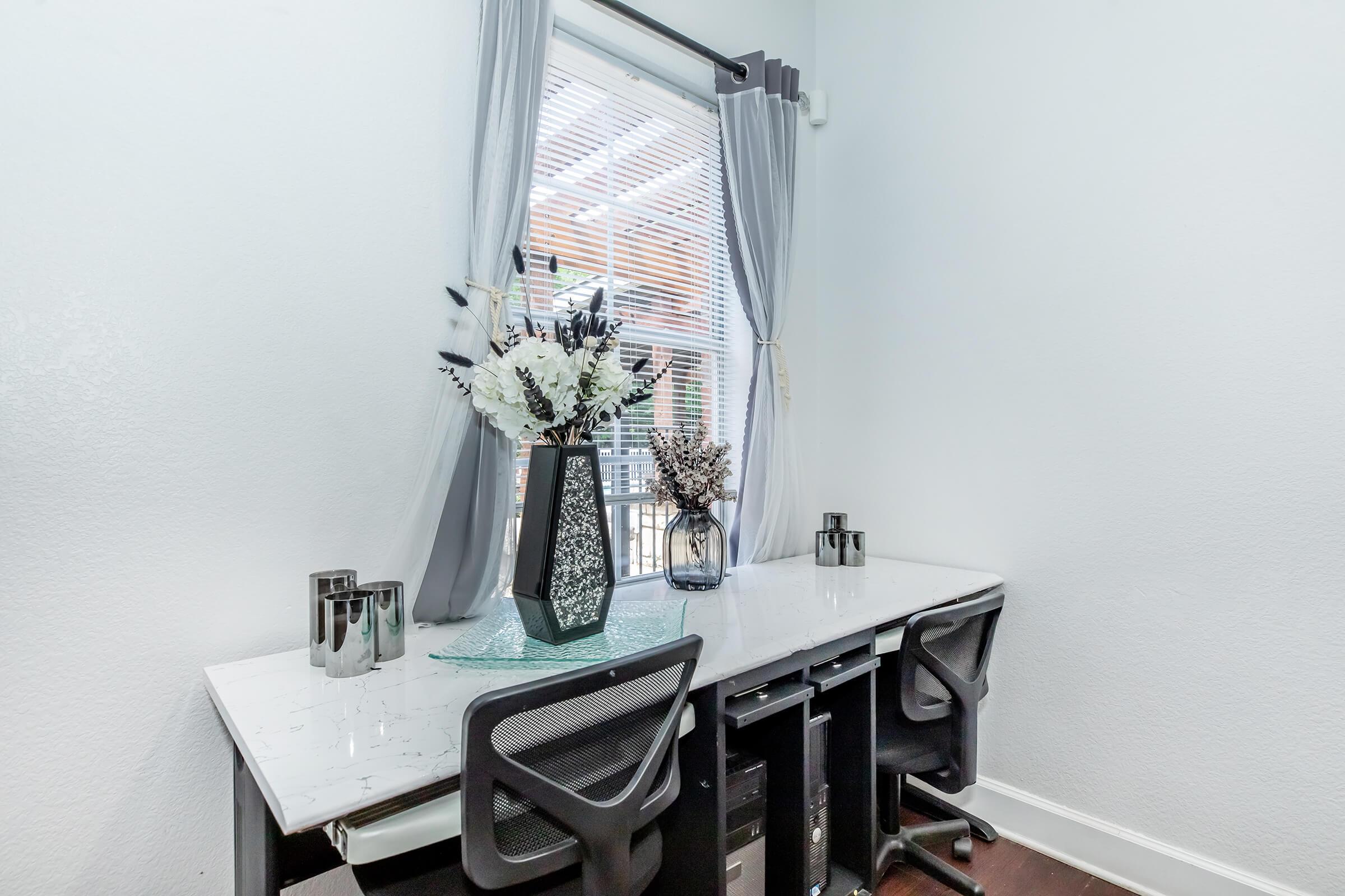 A modern home office setup featuring a white desk with two black chairs, adorned with vases of flowers. Natural light streams in through a window with sheer curtains. The wall is painted a light color, enhancing the airy feel of the space.