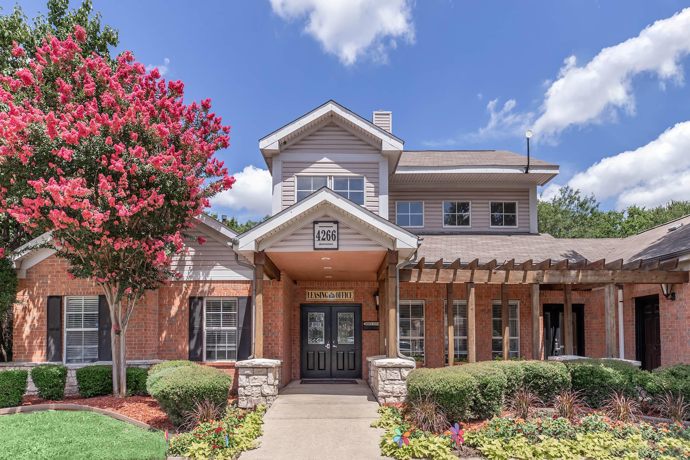 Exterior view of a two-story residential building with a welcoming entrance. The façade features red brick and tan siding, with decorative columns. Flanking the entrance are well-maintained flowerbeds with colorful plants. In the foreground, a flowering tree adds vibrant pink blooms against a blue sky with fluffy white clouds.