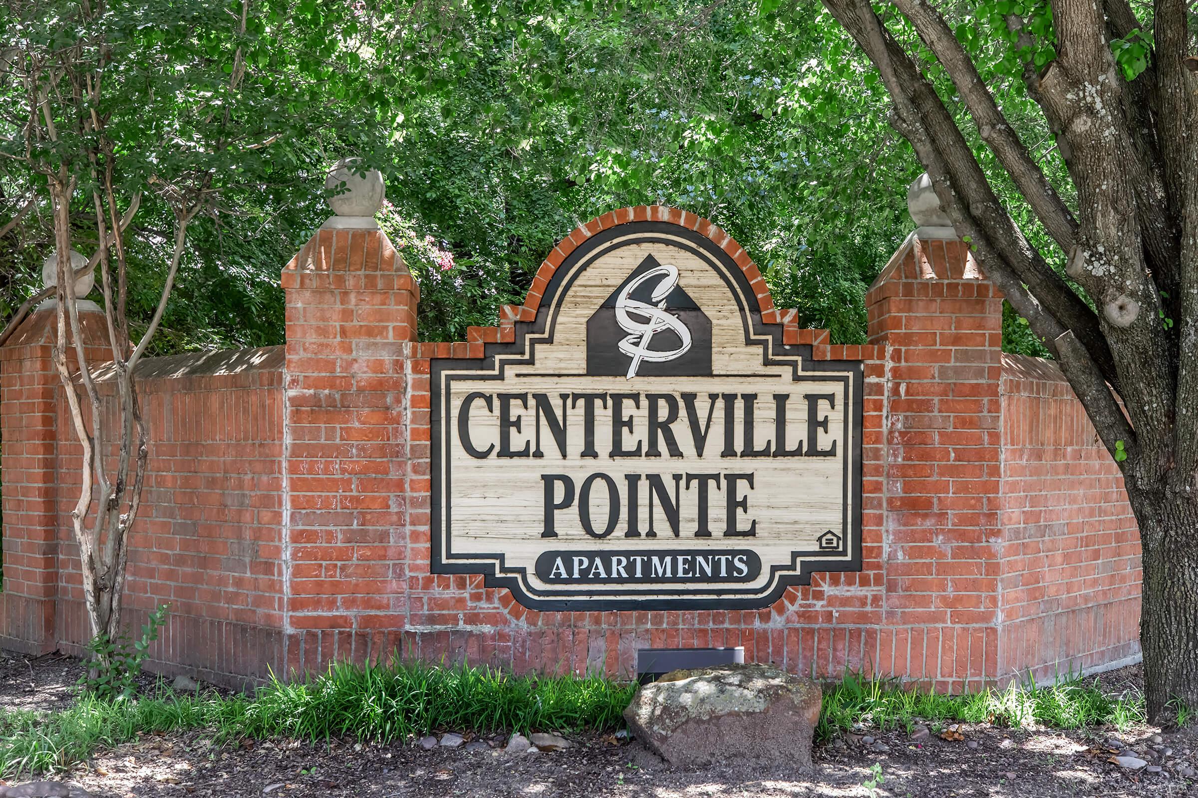 Sign for Centerville Pointe Apartments, featuring a wooden design with ornate details, surrounded by greenery and brick walls. The sign includes the name "CENTERVILLE POINTE" prominently displayed in large letters.