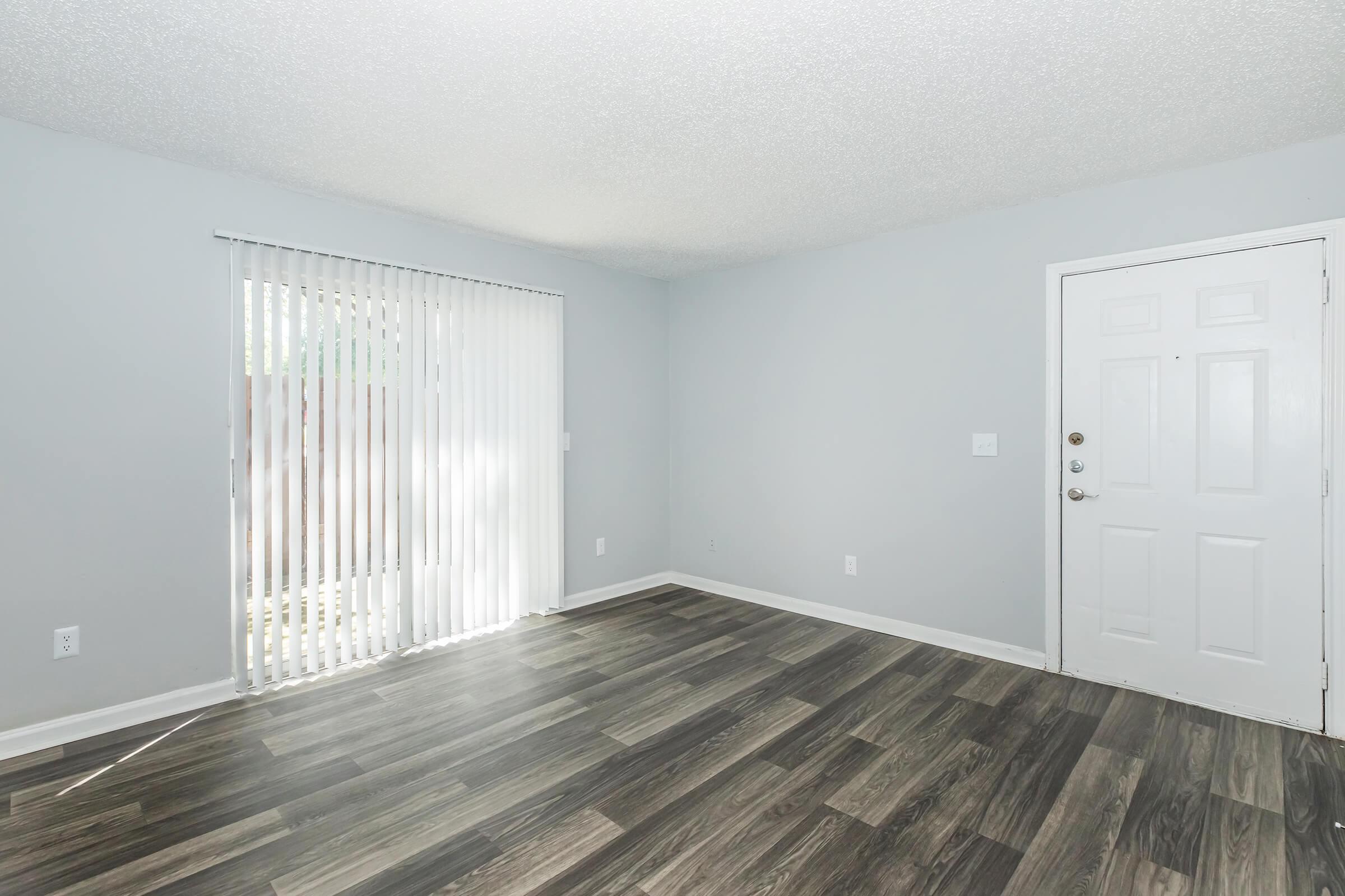 A spacious, unfurnished room with gray walls and a large window featuring vertical blinds. The floor has dark laminate or wood-like planks. A white door is visible on the right, providing access to the outside. The overall ambiance is bright and airy, perfect for personal customization.