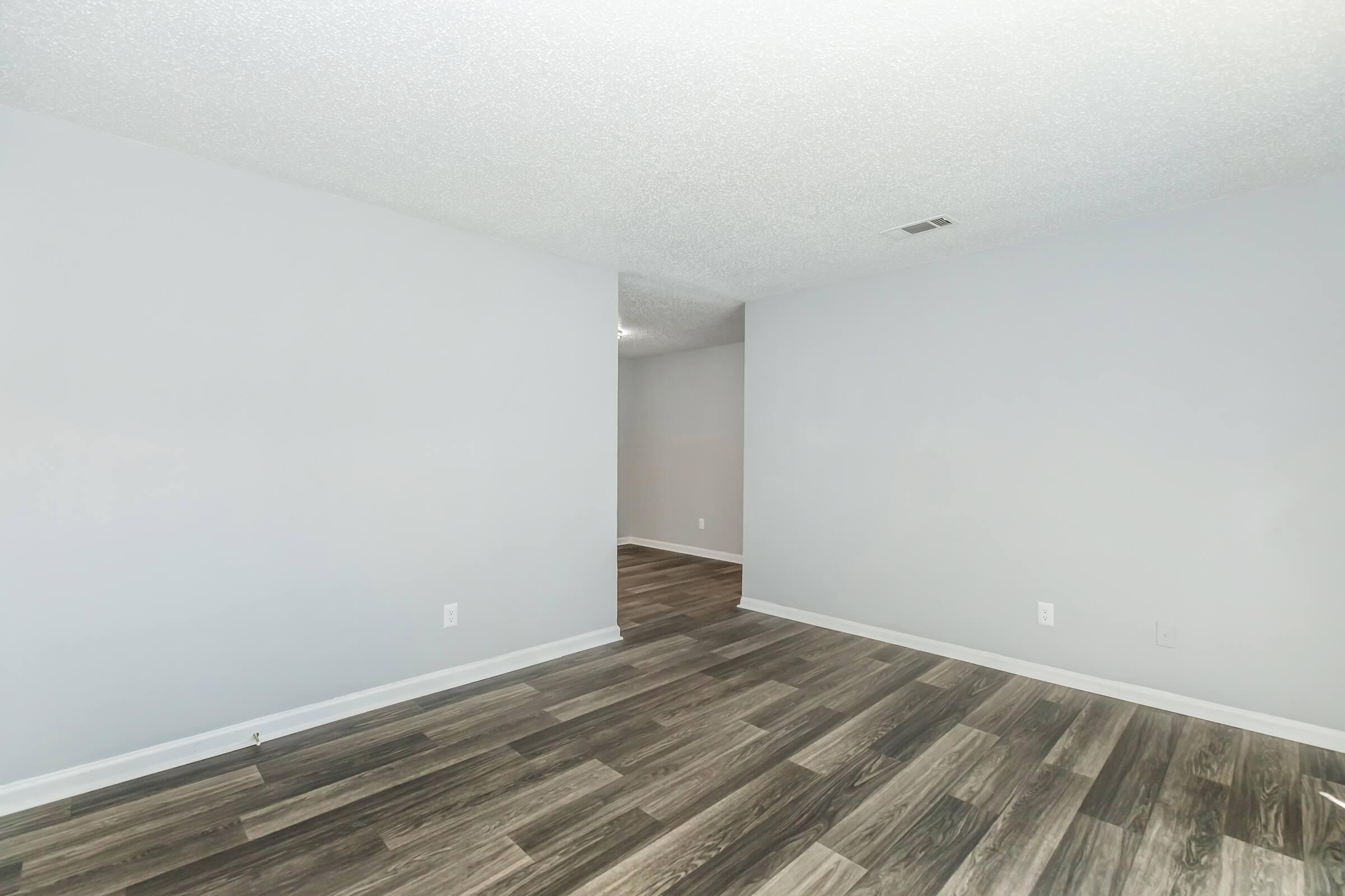 A spacious, empty room with light gray walls and a textured ceiling. The floor features dark wood-like laminate planks. A doorway leads to another room, and there is ample natural light. The overall ambiance is modern and minimalist, suitable for various design possibilities.