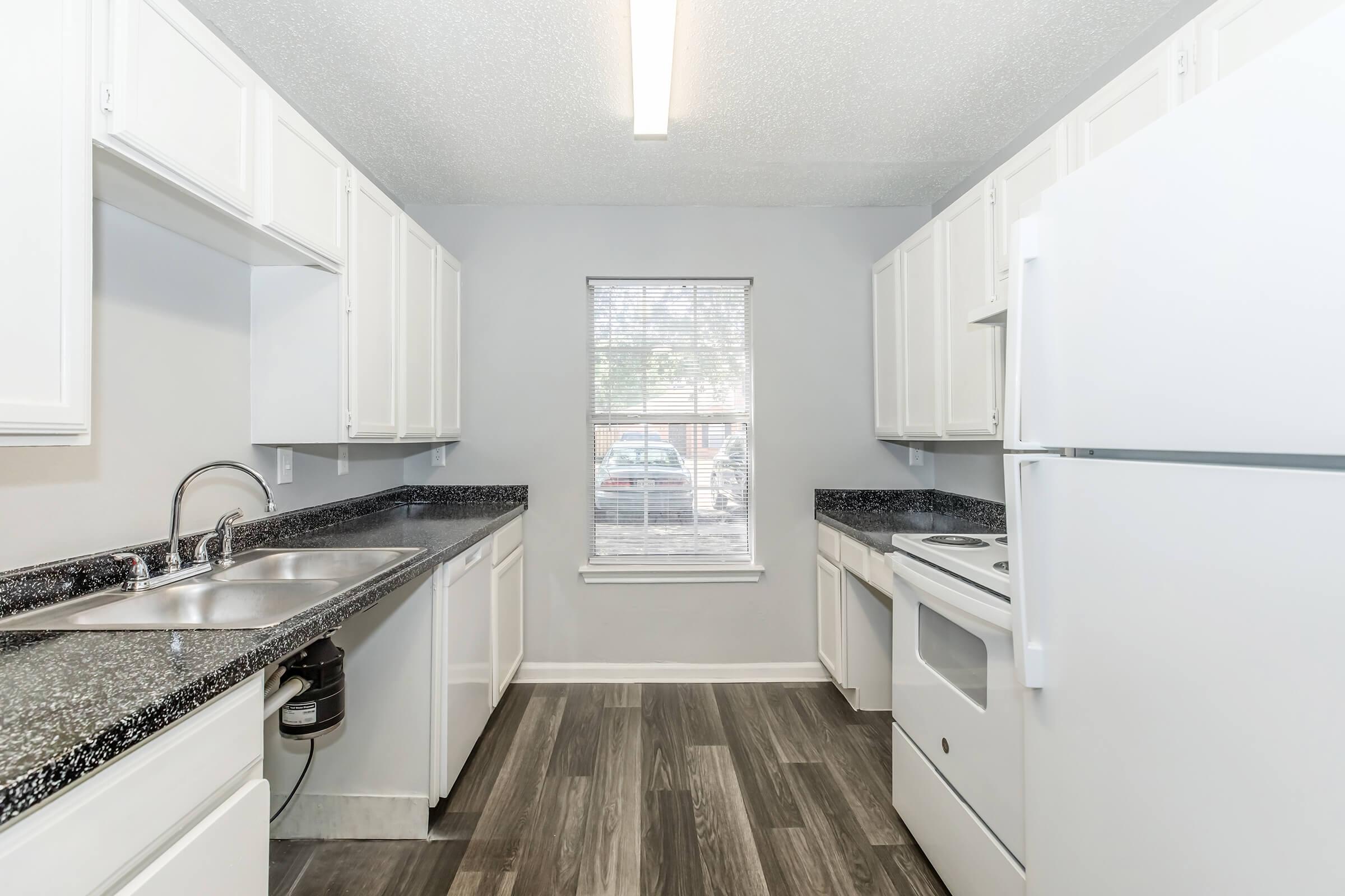 A modern kitchen featuring white cabinets, a double sink with a garbage disposal, and a stove with an oven. The countertop is dark and speckled. Natural light comes in from a window, illuminating the room. Wood-look flooring completes the contemporary design.