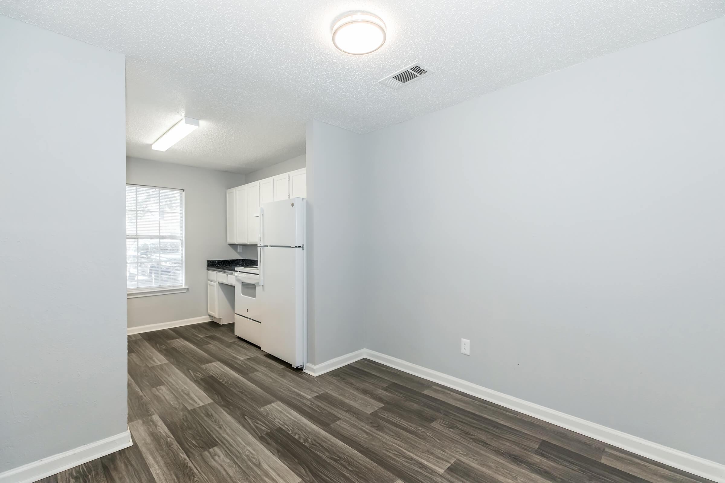 A modern kitchen space featuring light gray walls, white cabinetry, and a black countertop. A refrigerator is visible, and there's ample natural light coming from a window. The floor is made of dark wood-style laminate, contributing to a clean, minimalist aesthetic.