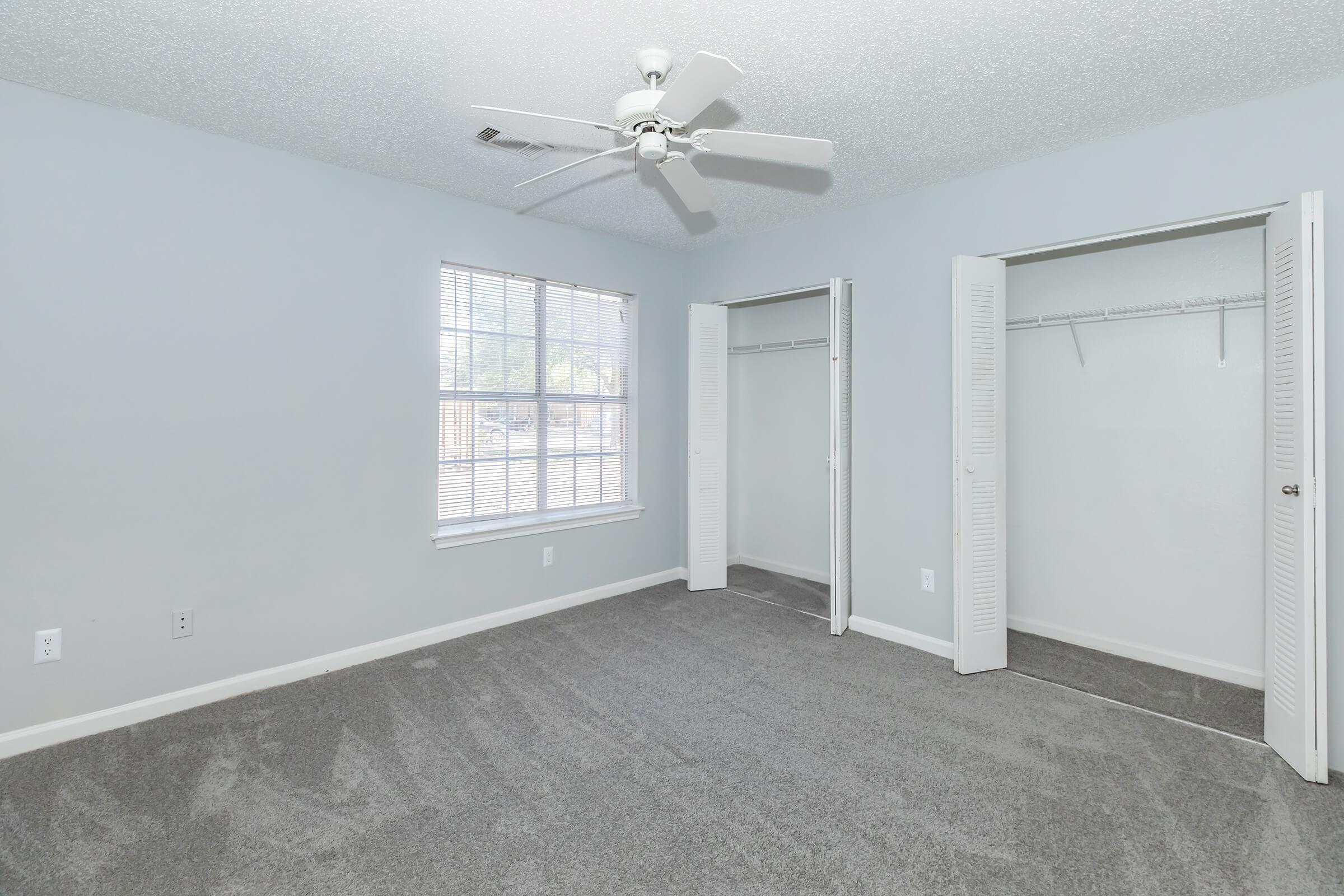 Empty bedroom featuring light gray walls, a ceiling fan, large window with blinds, and plush gray carpet. There are two built-in closets with sliding doors, providing ample storage space. The room is well-lit and inviting, ideal for personalizing or moving in.