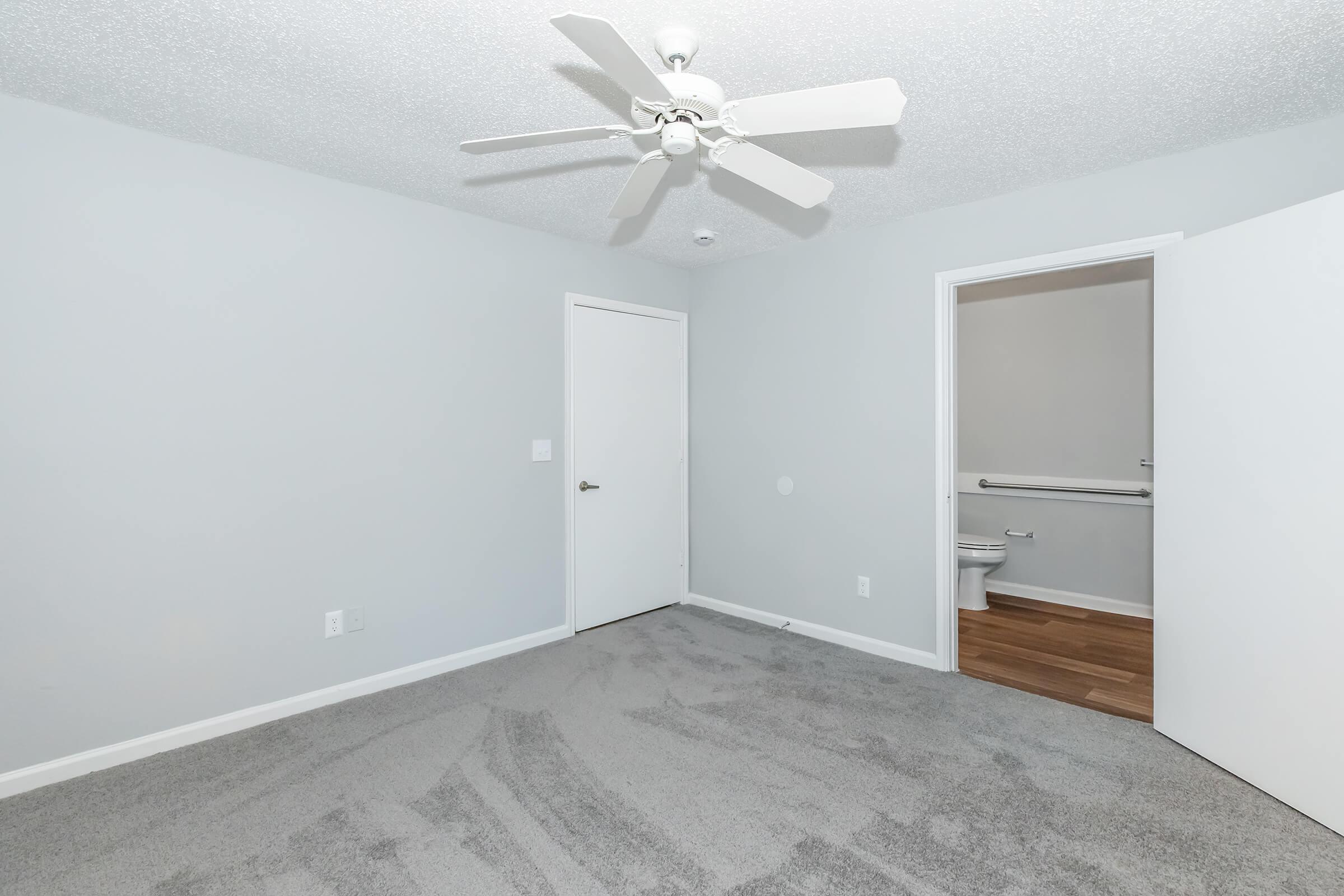 A spacious, light gray bedroom featuring a ceiling fan, freshly carpeted flooring, and a neutral color palette. There is a white door leading to a bathroom visible in the background, providing a clean and tidy atmosphere.