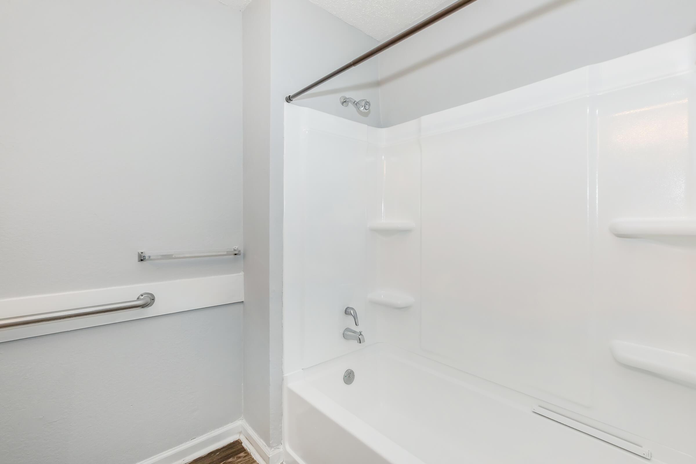 A clean, modern bathroom featuring a white tub with a shower curtain, grab bars for safety, and smooth gray walls. The space is well-lit with an open design, making it feel spacious and accessible.
