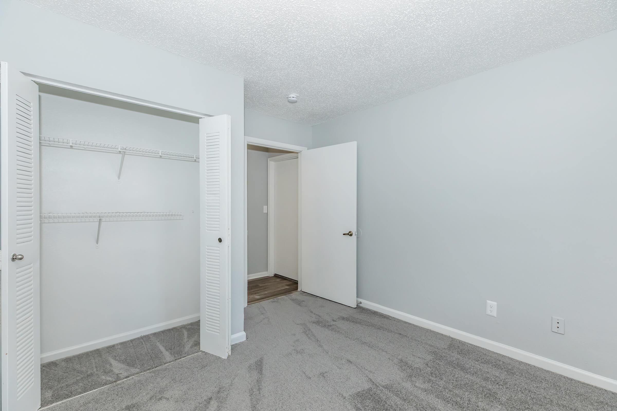 A small, empty room with light gray walls and carpet. It features a closet with open doors, showcasing hanging shelves. On the right, there's a closed door leading to another area. The room has a clean and modern appearance, with natural light filtering in.