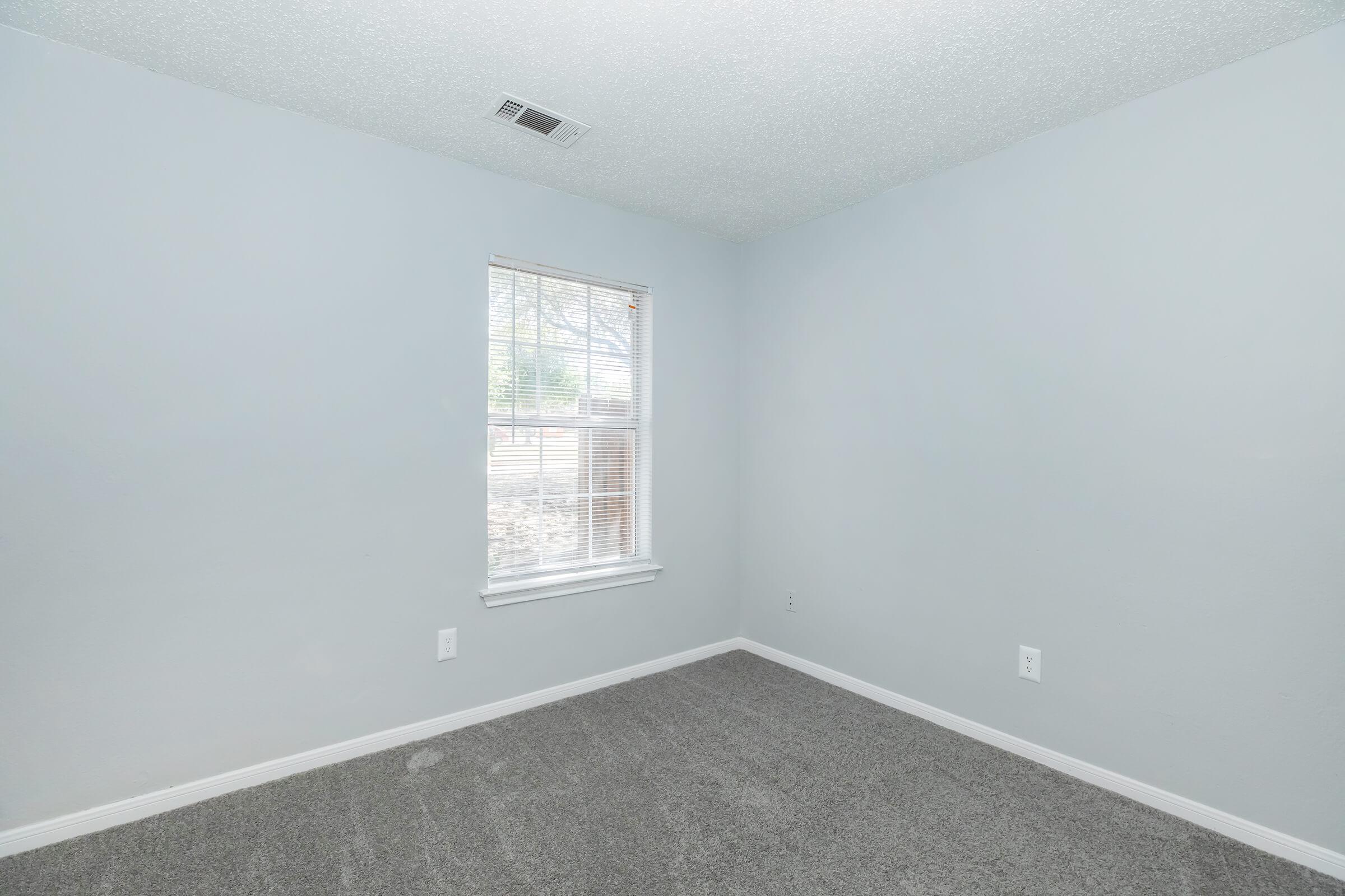 Empty room with light gray walls and carpet. A single window allows natural light to enter, featuring white blinds. The space appears clean and minimalistic, suitable for various uses such as a bedroom or office.