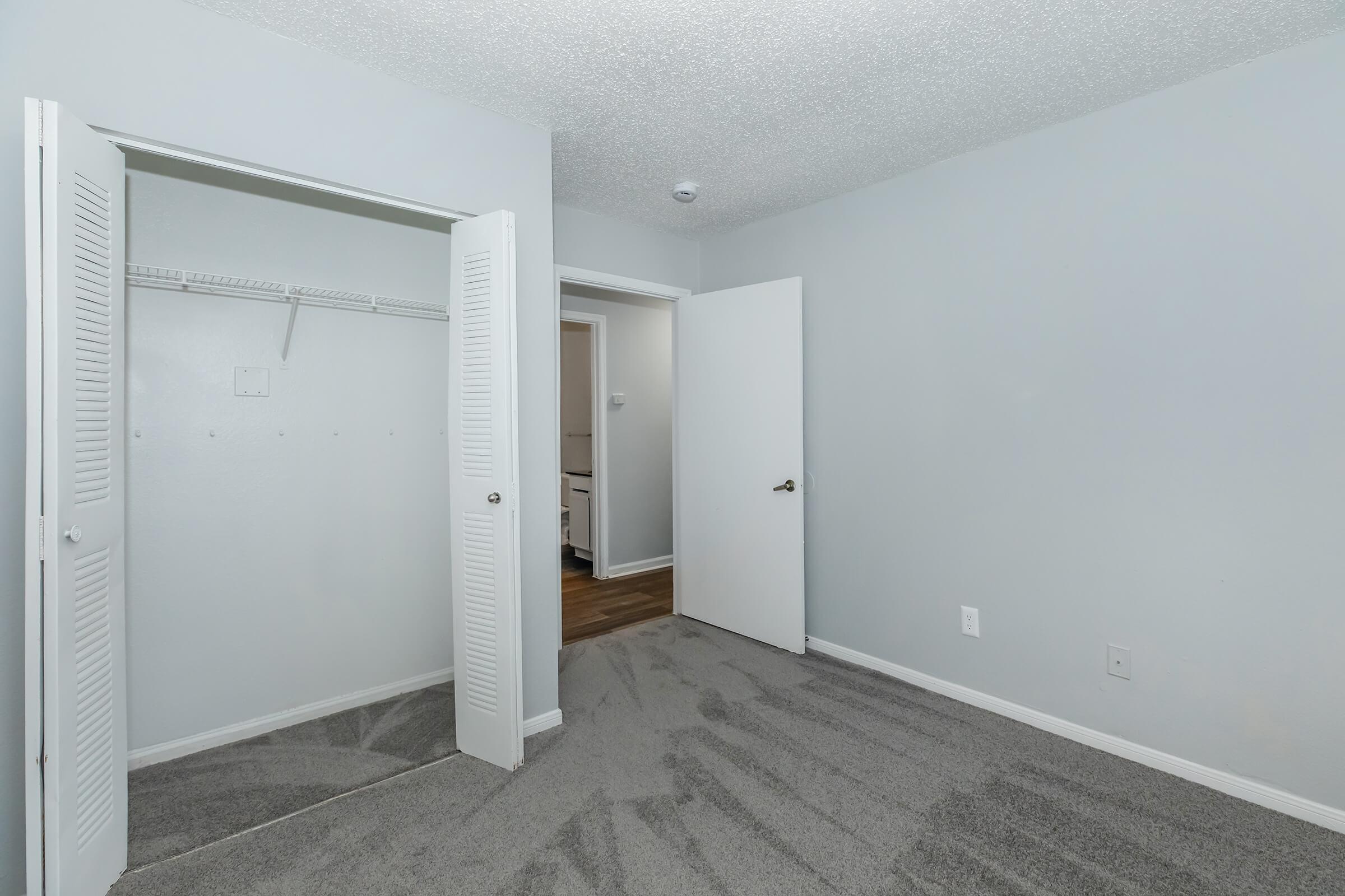 A vacant room with light gray walls and carpet. There are two white doors: one leads to a closet with shelves, and the other is an entryway. The room has no furniture and appears clean and well-lit, with a ceiling texture visible.