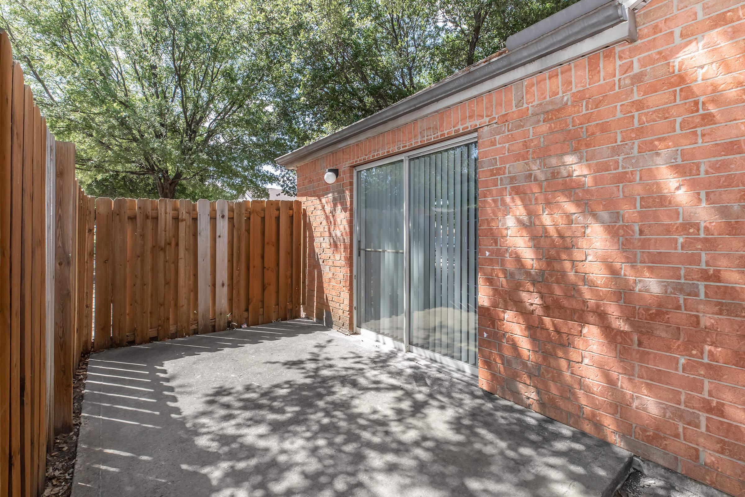 A small outdoor patio area featuring a concrete floor and a sliding glass door surrounded by a brick wall and wooden fence. Sunlight filters through the trees, casting shadows on the ground, creating a tranquil and private space.