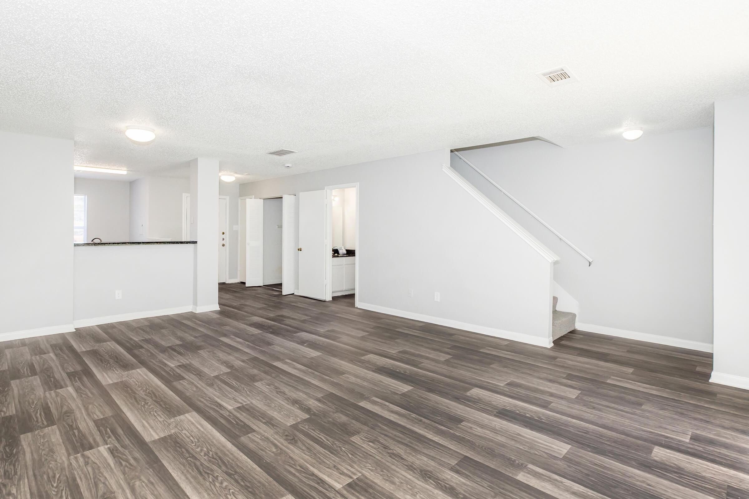 Spacious interior of a modern living area featuring light gray walls, textured ceiling, and wood-style flooring. The layout includes an open space leading to a staircase, with doorways leading to other rooms in the background. Natural light fills the room, enhancing the airy feel.