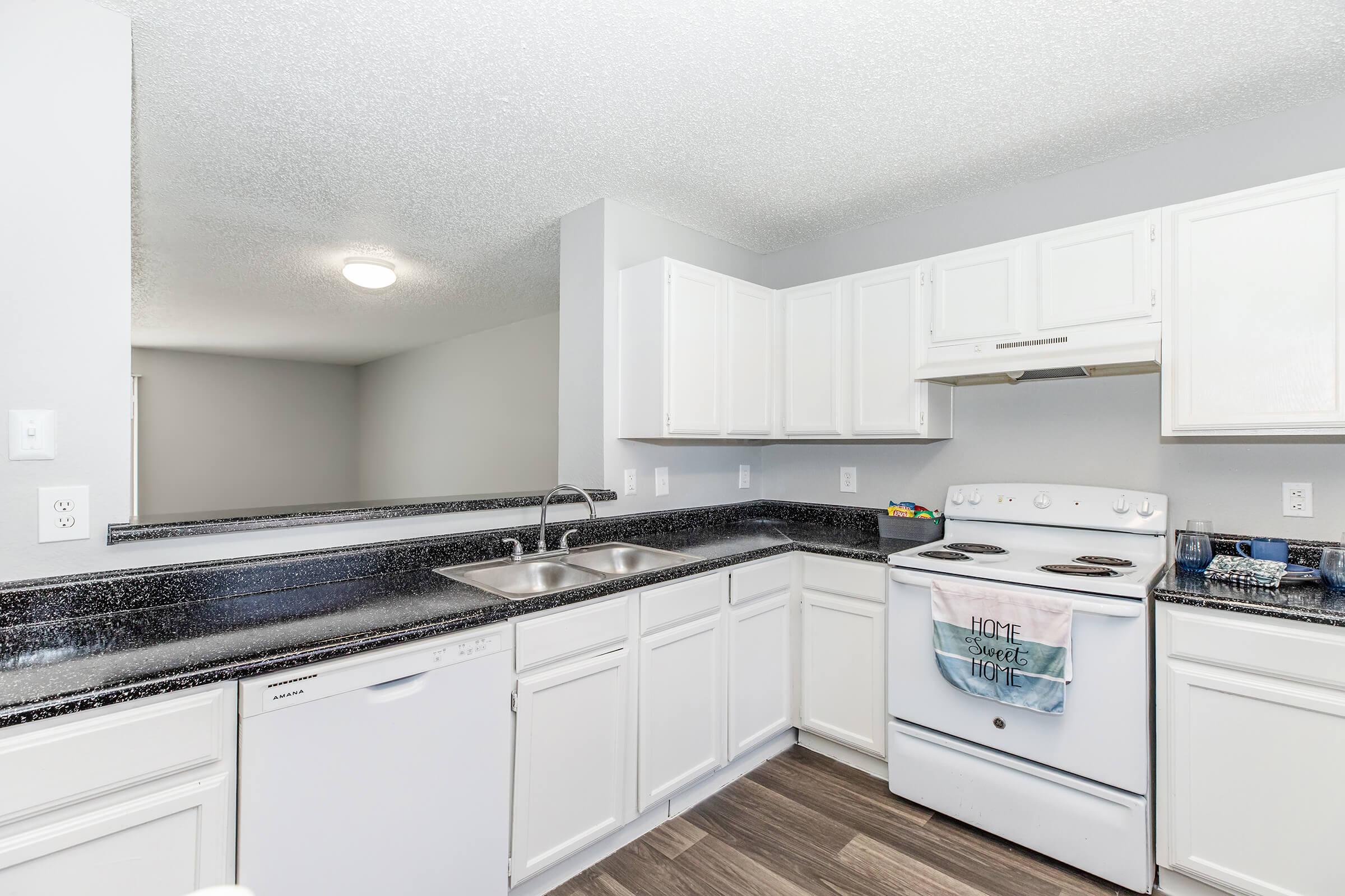 Modern kitchen featuring white cabinets, a black granite countertop, a double sink, and a white stove with a hood. Appliances include a dishwasher. The space has a neutral color palette and wood-like flooring, with a bright overhead light. A separate room is visible in the background.