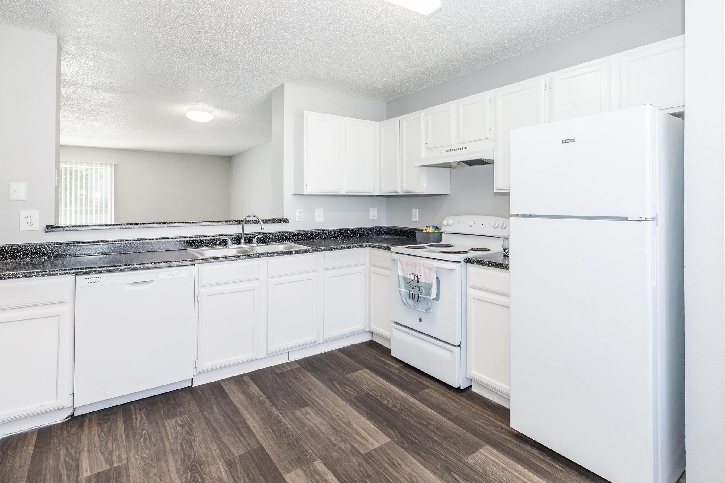 Bright and modern kitchen with white cabinetry, black countertops, and a full set of appliances including a refrigerator, stove, and dishwasher. The flooring is dark wood laminate, and there's ample natural light coming through a nearby window.