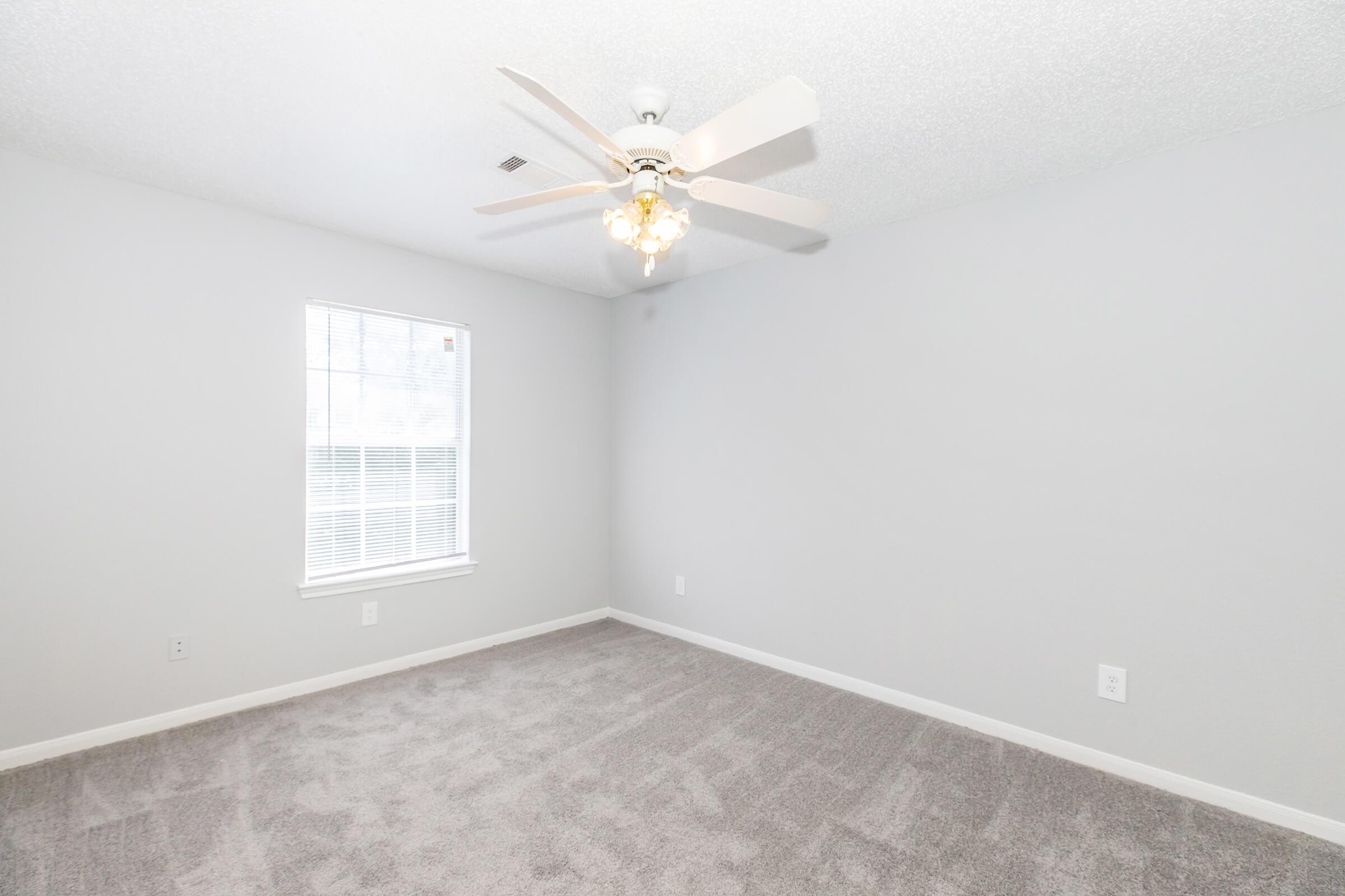 A spacious, empty room with light gray walls and plush carpet. It features a ceiling fan with a light fixture and a window allowing natural light to enter. The room has a minimalistic design, creating a bright and airy atmosphere suitable for various purposes.
