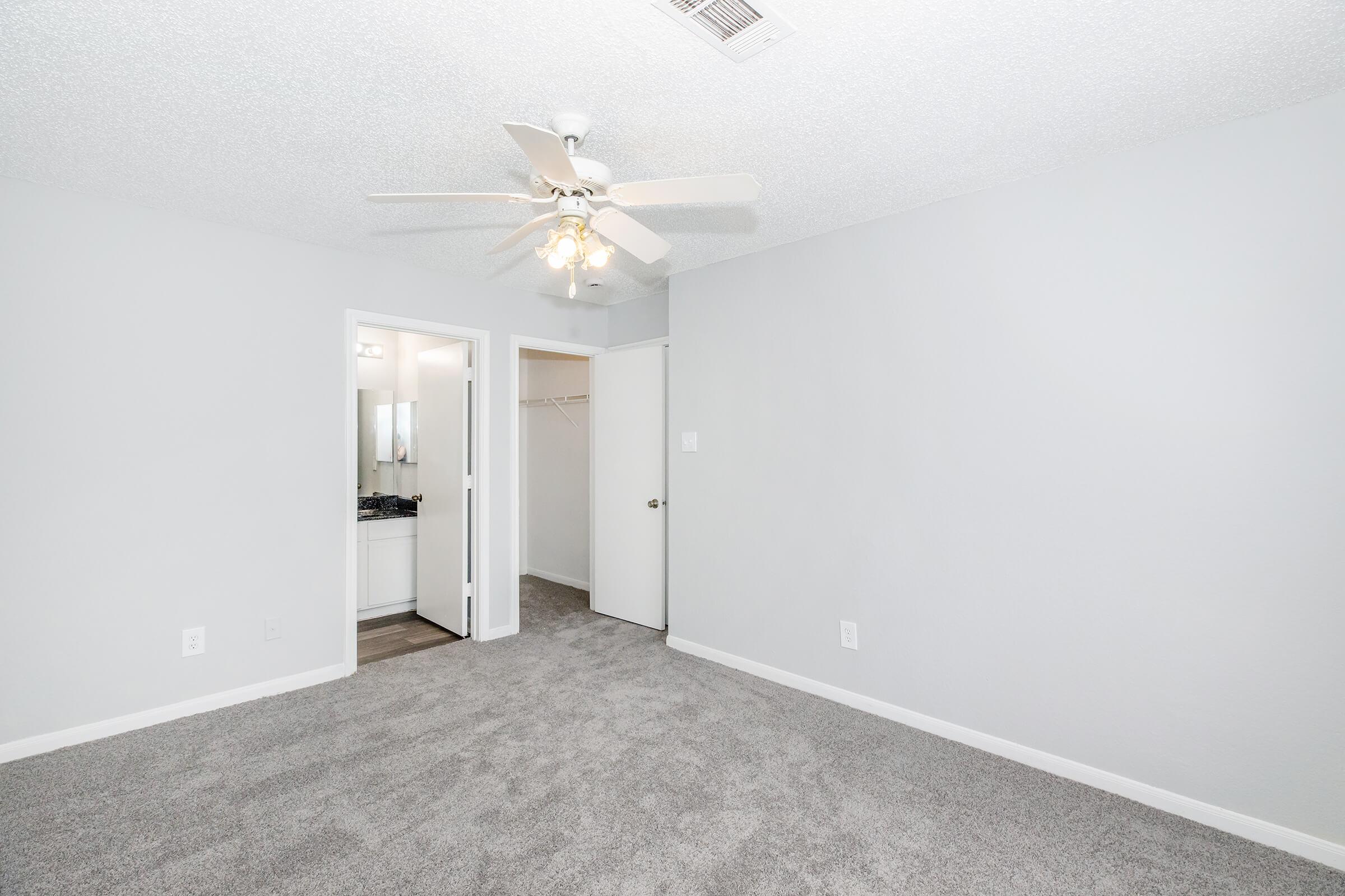 A bright, carpeted bedroom with light gray walls. A ceiling fan is installed, and there are two doorways: one leading to a small bathroom area with a mirror, and another leading to a closet. Natural light enters the room, creating a spacious and welcoming atmosphere.