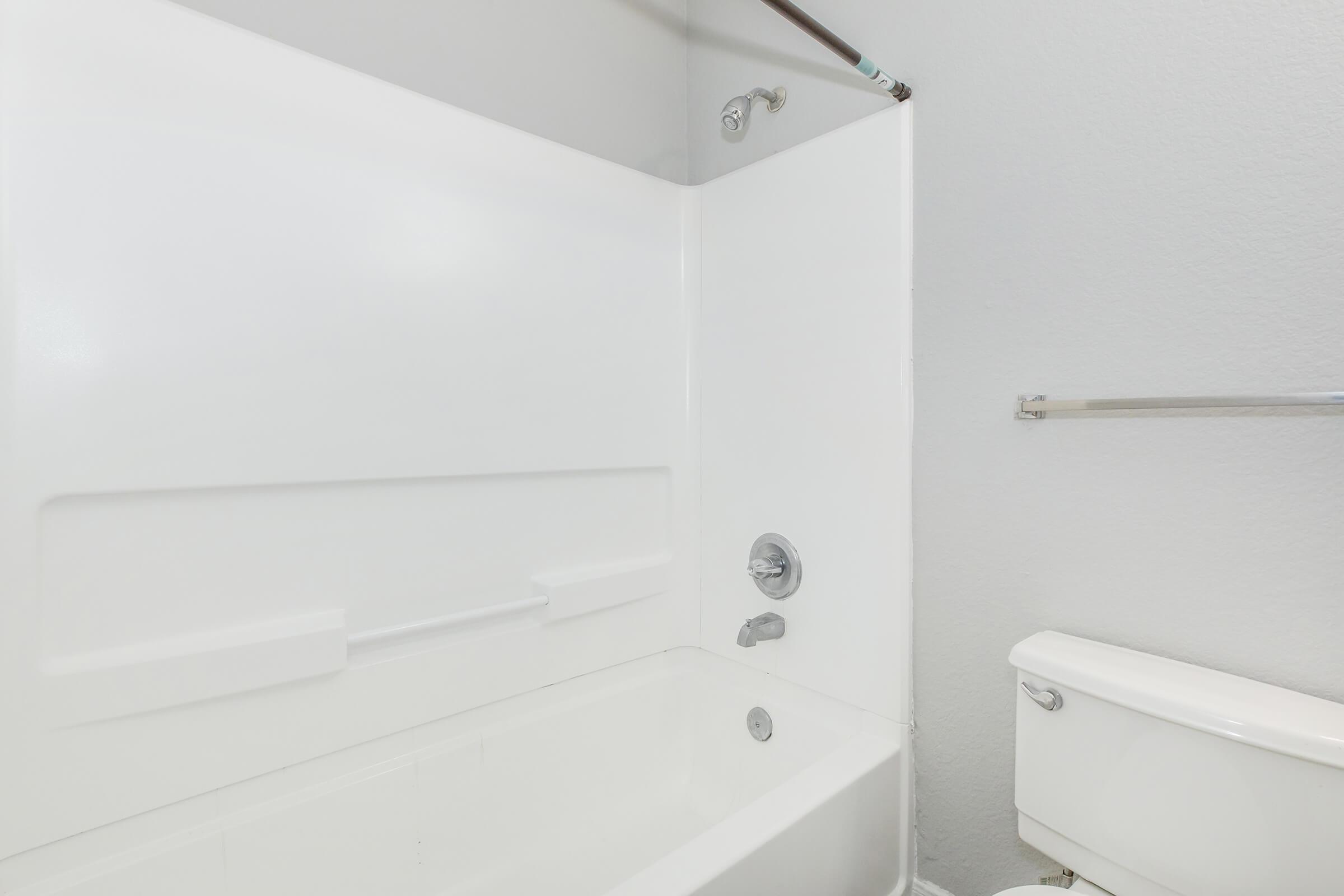 A simple bathroom featuring a white bathtub with a showerhead and a wall-mounted faucet. There is a toilet beside the bathtub, both fixtures set against plain light-colored walls. A towel rack is mounted on the wall near the toilet. The space appears clean and minimalistic.