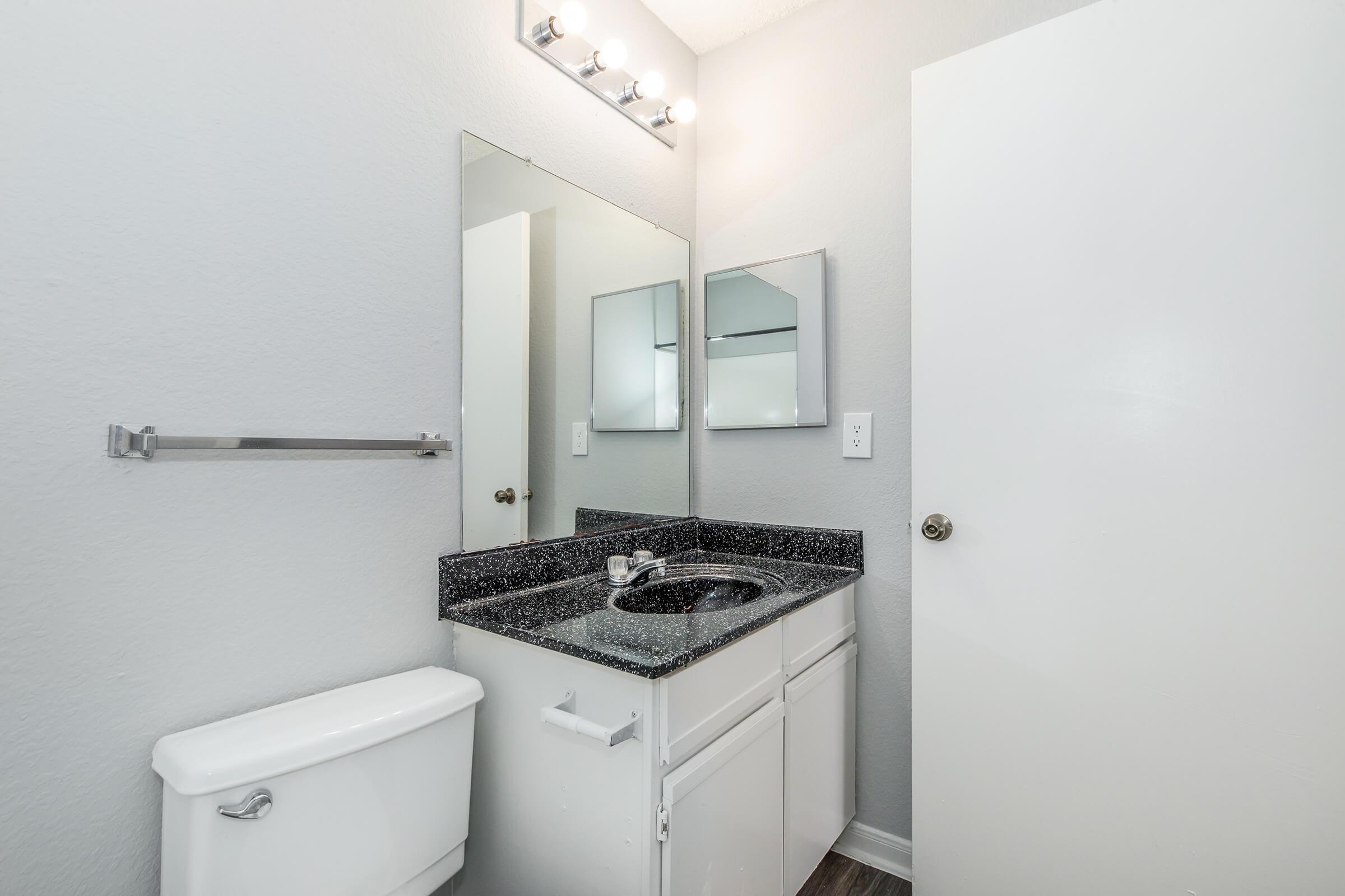 A modern bathroom featuring a dark granite countertop with a sink, a large mirror above, and two smaller mirrors on the wall. The walls are painted light gray, and there's a white toilet nearby. The space is well-lit by overhead lights.